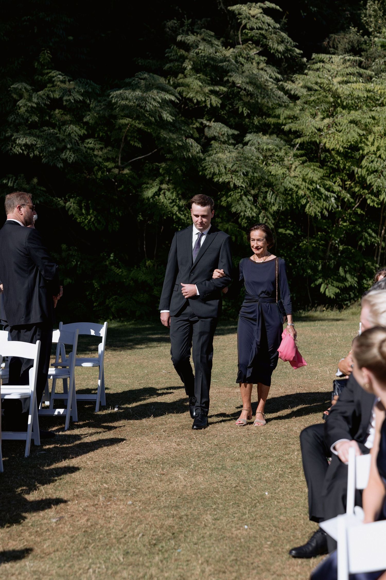 Wedding party members walk across a grass lawn with white chairs arranged for the ceremony.