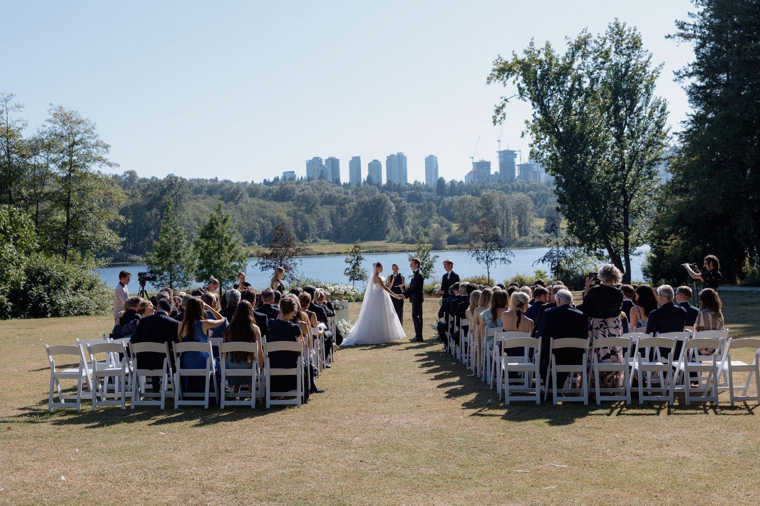 Panoramic view of outdoor wedding ceremony setup with white chairs and city skyline in background and Deer Lake at Hurt House in Burnaby