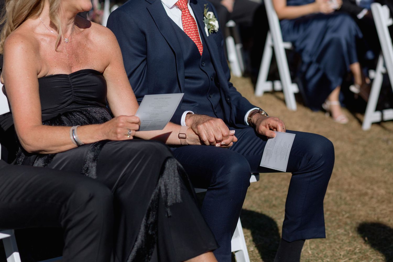 Wedding guests in black dresses and suits hold ceremony programs during an outdoor service.
