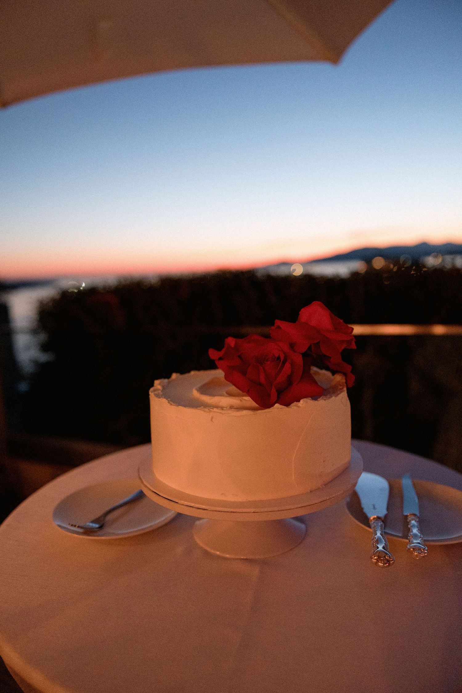 White wedding cake topped with red roses on an outdoor table at sunset.