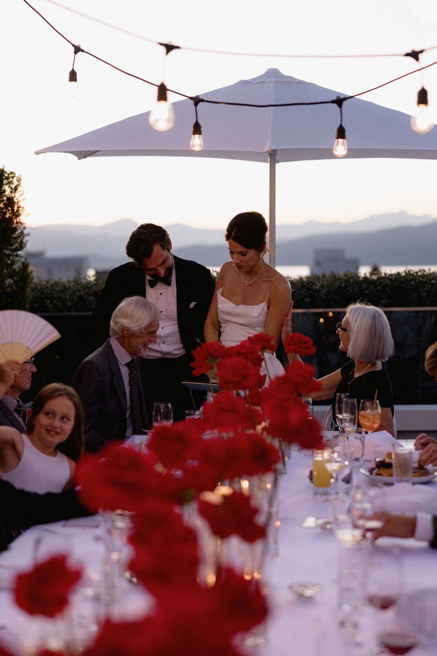Outdoor wedding reception at sunset with string lights overhead and vibrant red flowers on white tablecloths.