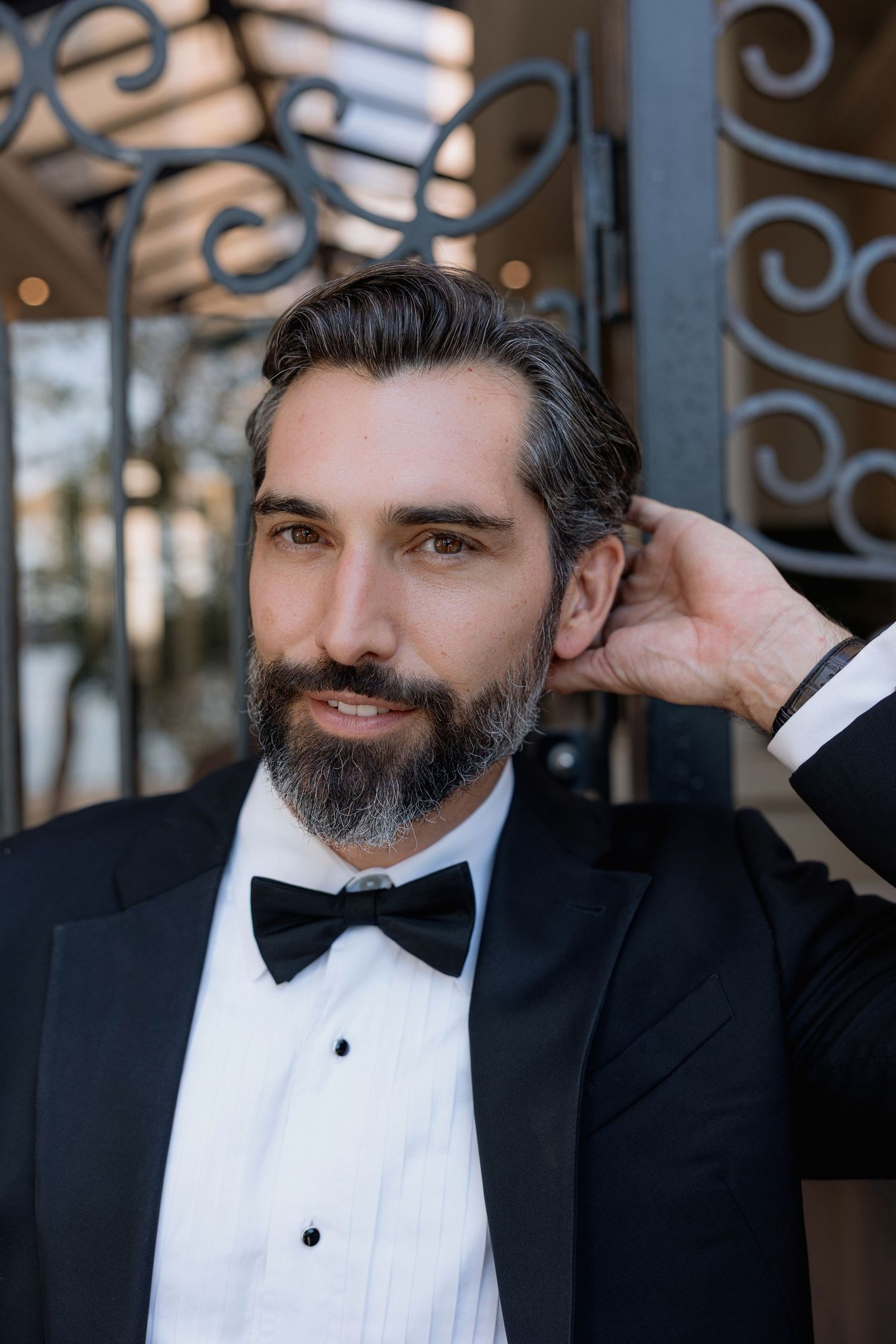 A well-dressed man in a black tuxedo and bow tie poses elegantly against an ornate iron gate.