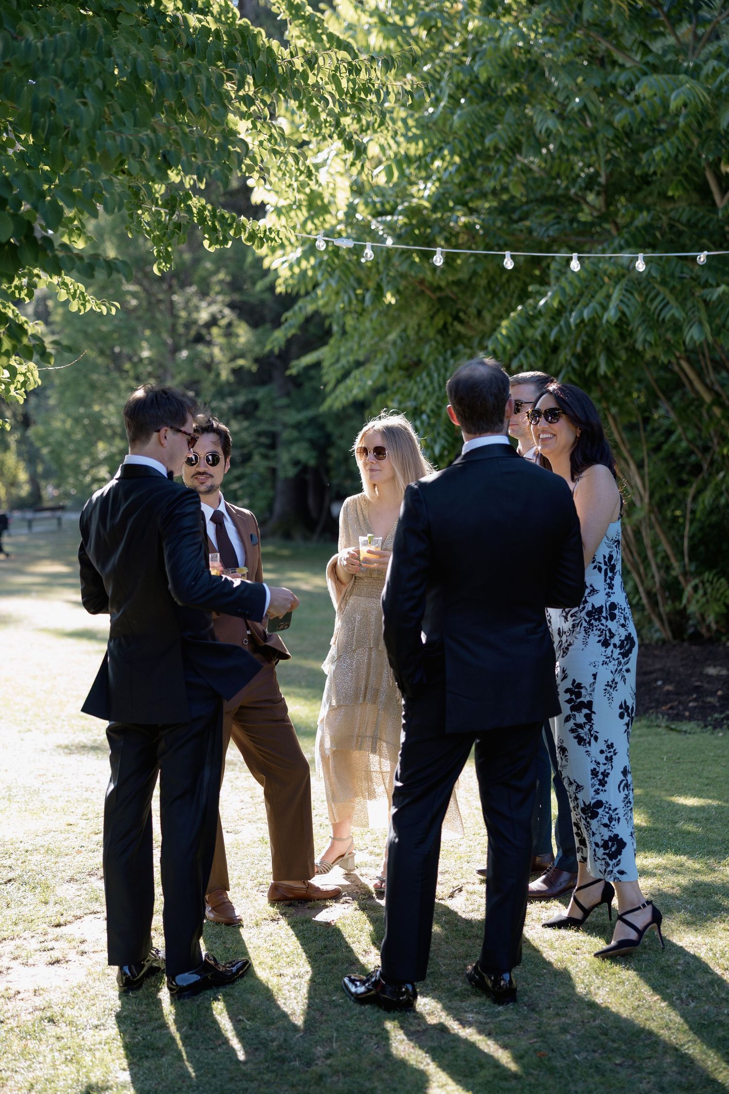 Small group of wedding guests chatting in formal attire under string lights at outdoor reception.