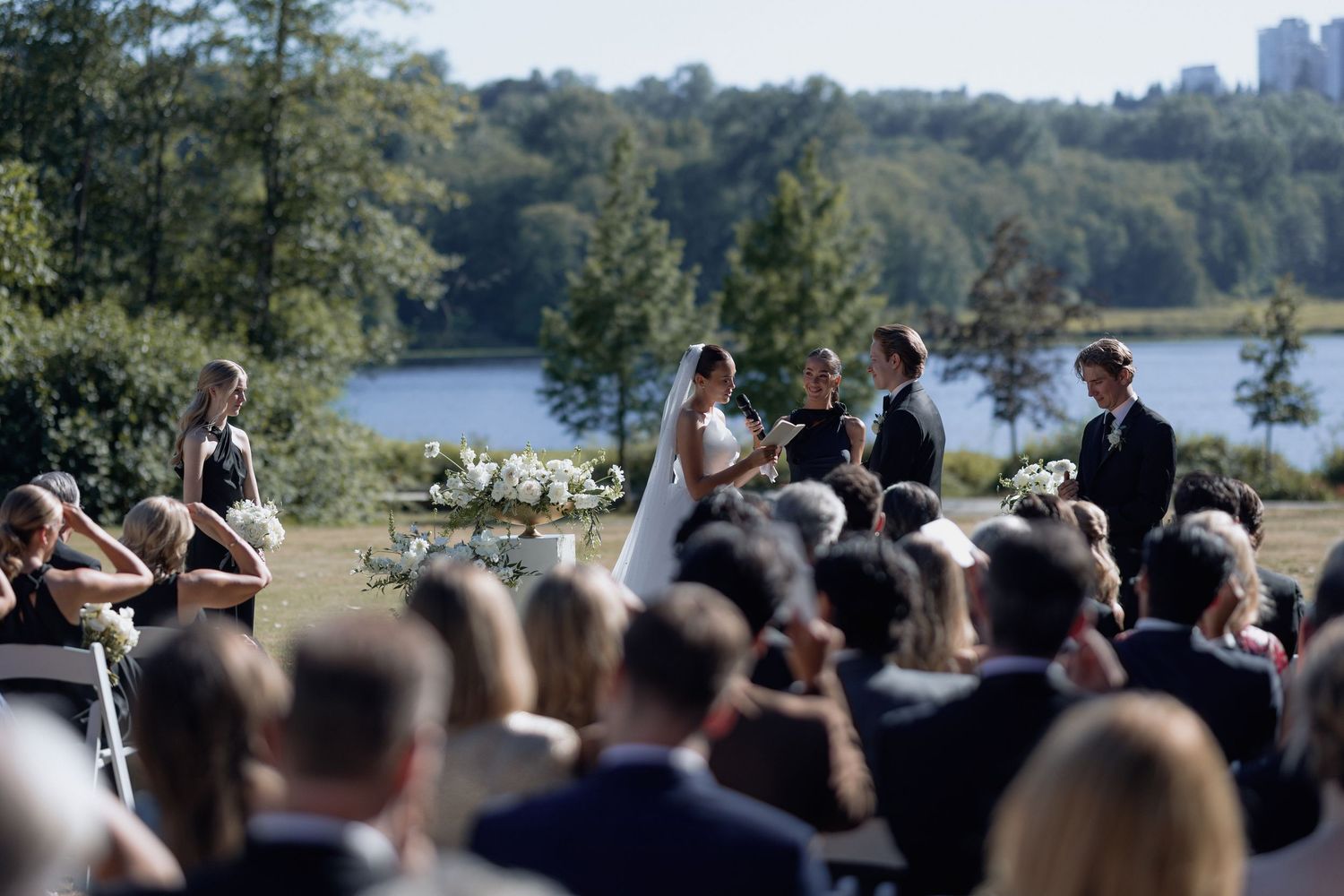 Panoramic view of wedding guests gathered for an outdoor ceremony with a lake and urban skyline in the distance.