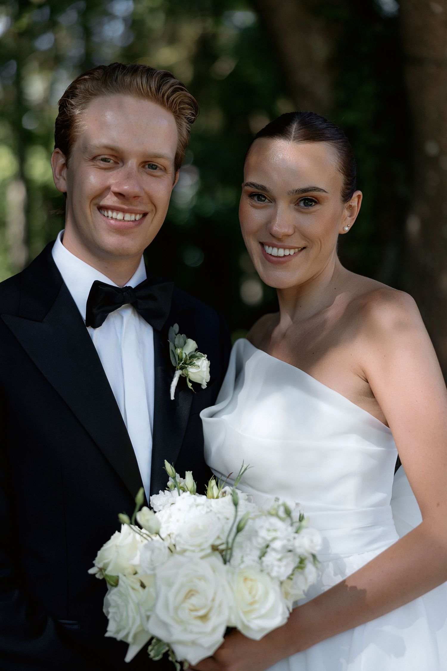 Elegant wedding couple with white rose bouquet poses in a forest setting.