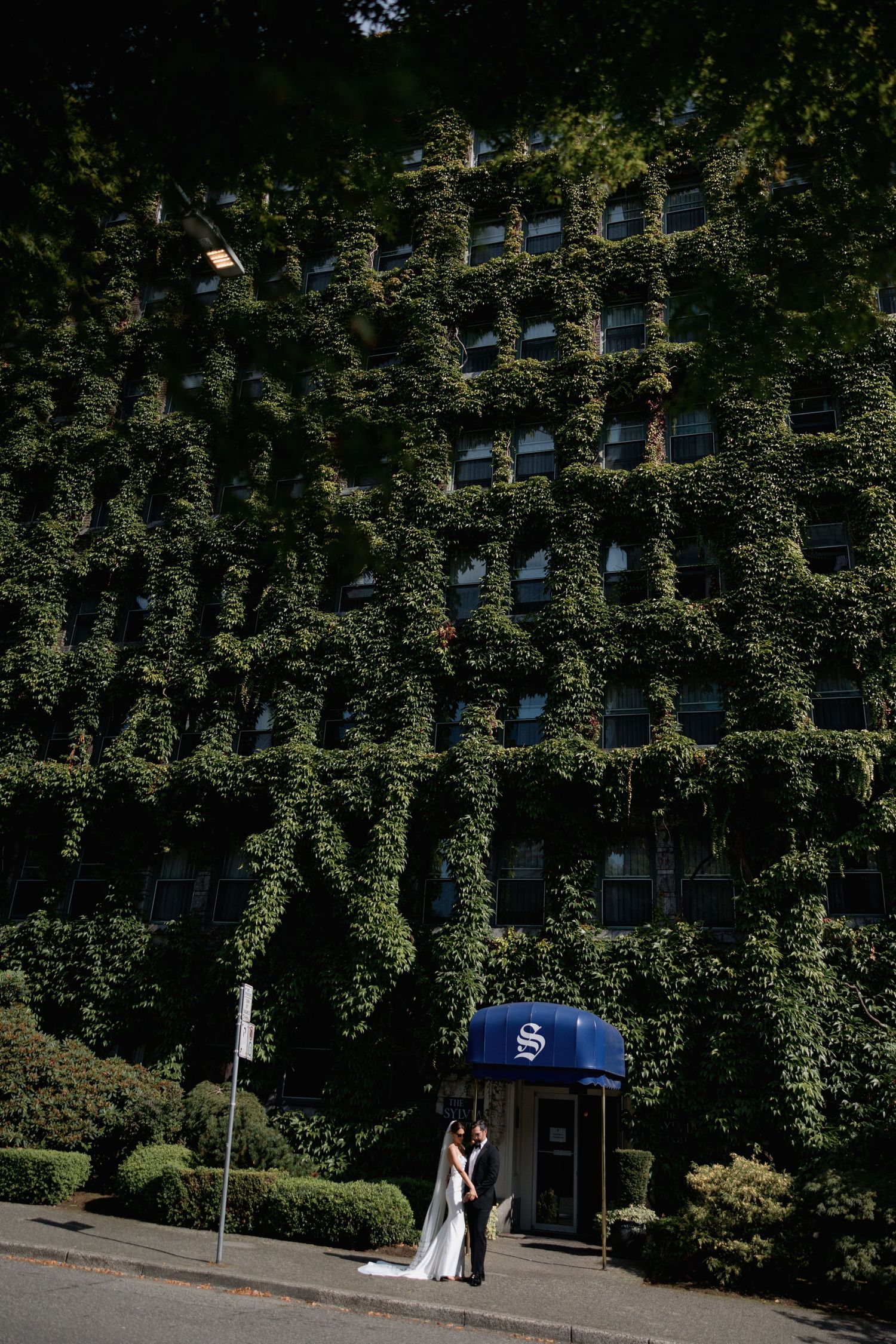 A wedding couple stands outside an ivy-covered building entrance with blue awning in front of the Sylvia Hotel in Vancouver