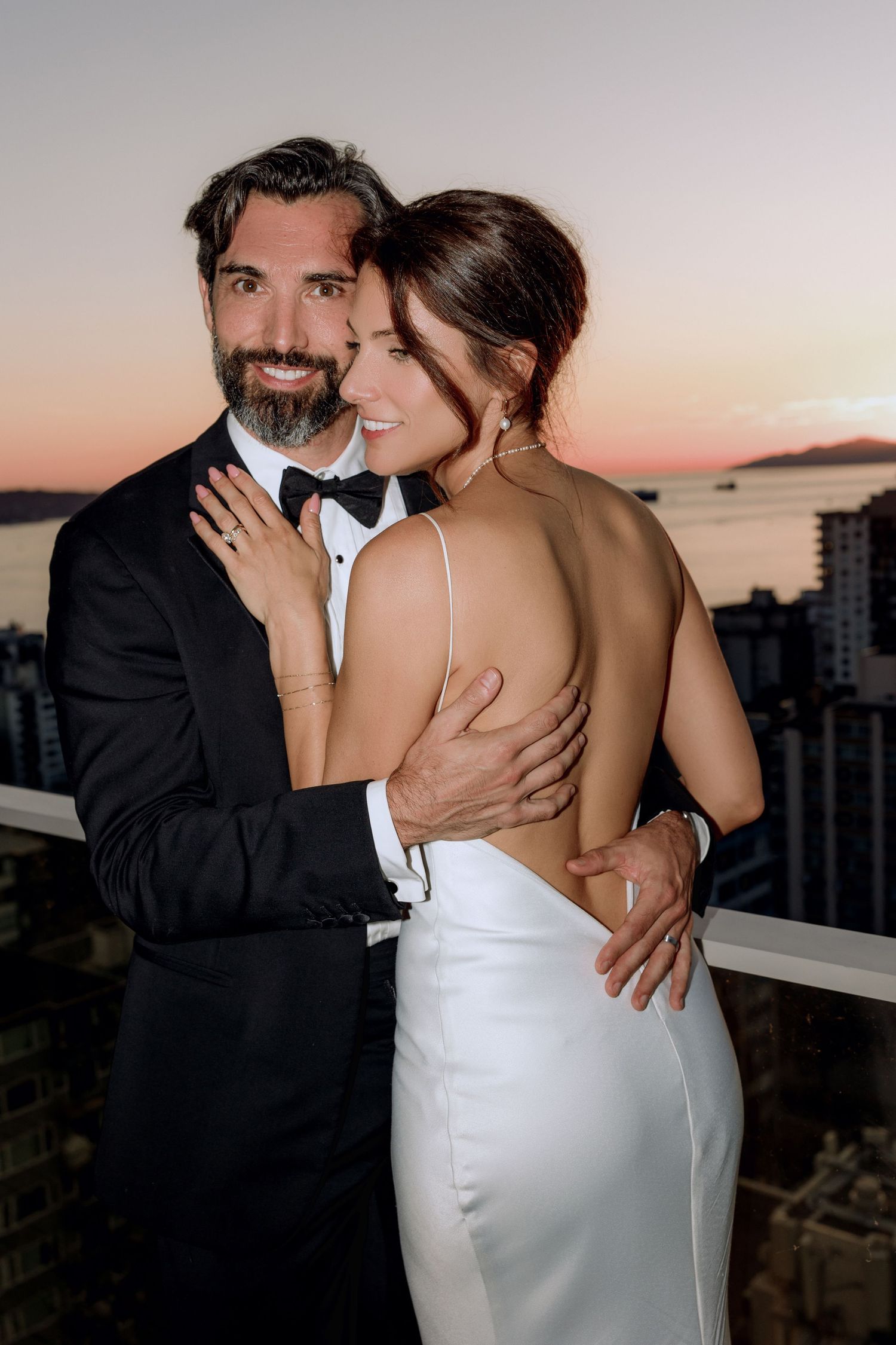 Romantic couple embrace at sunset on rooftop terrace in formal attire.
