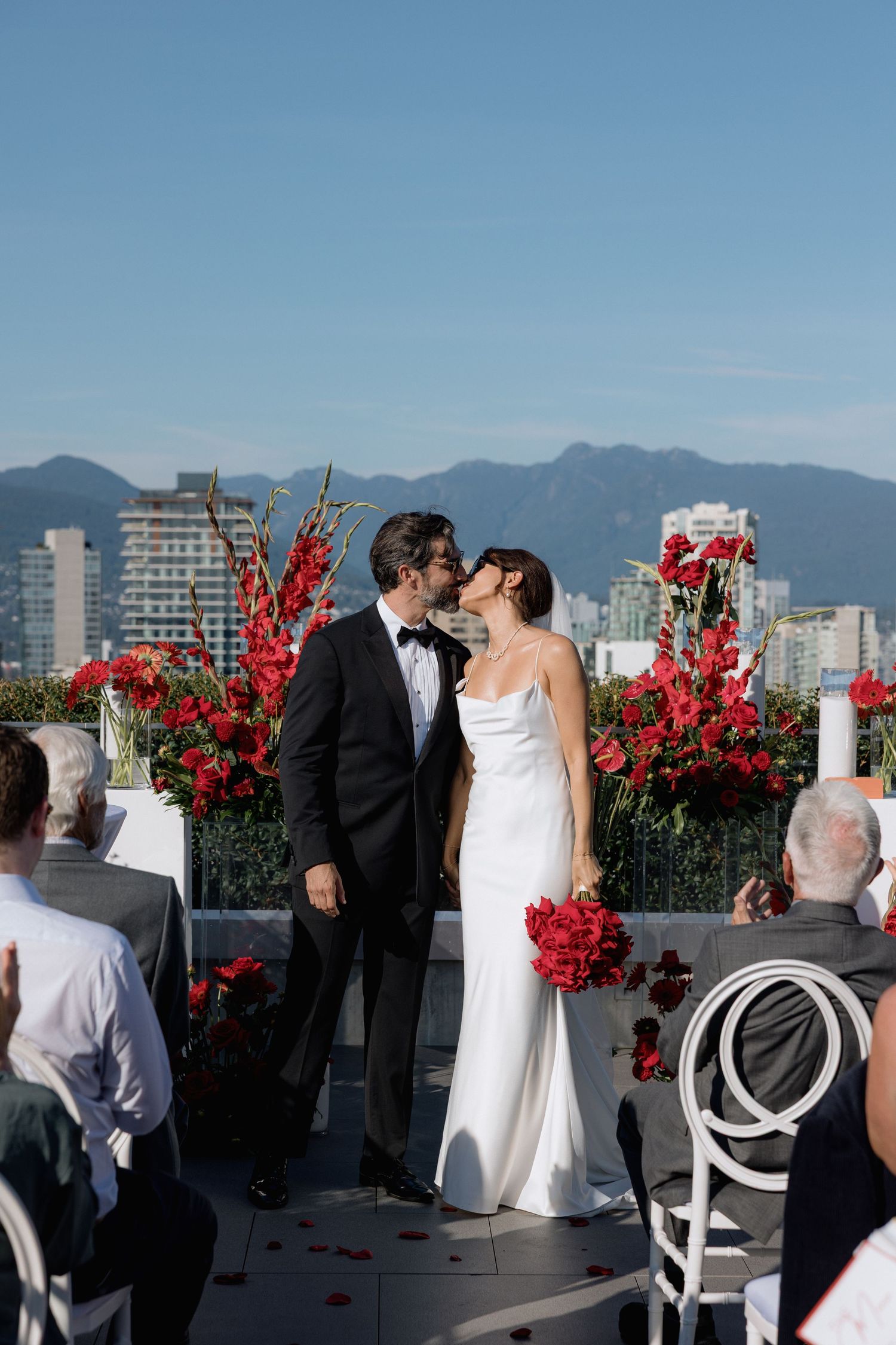 Outdoor rooftop wedding ceremony with bright red floral arrangements and city skyline backdrop.