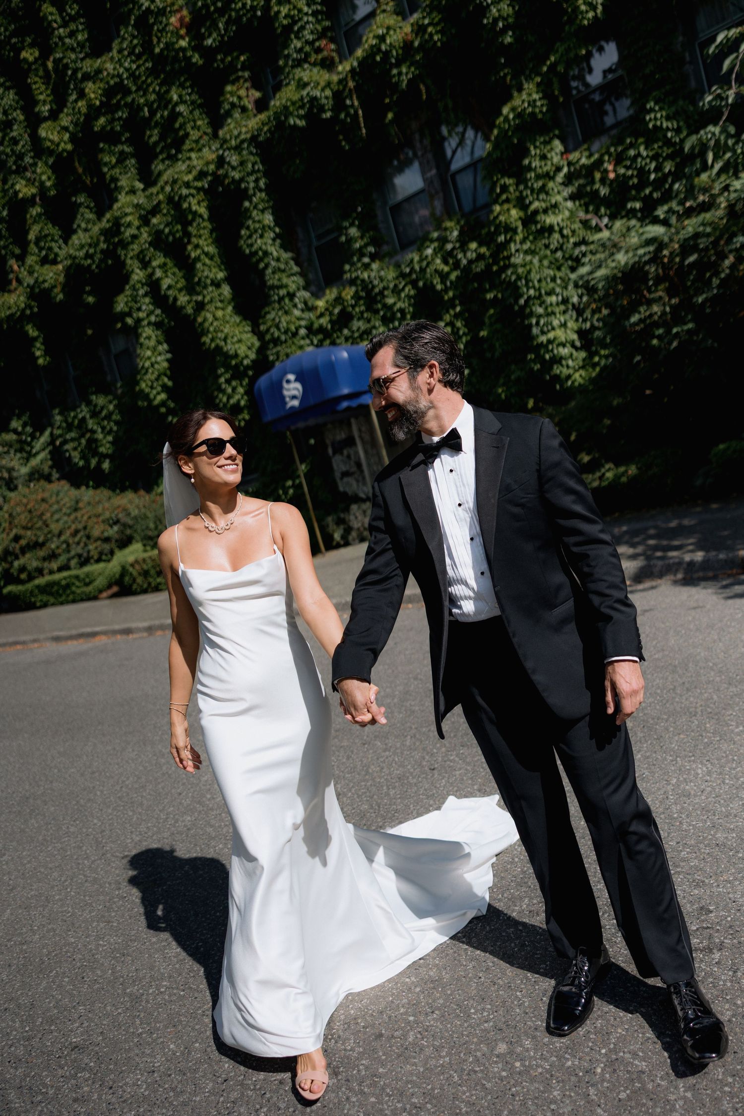 Newlyweds walking hand in hand on a sunny day with greenery and a blue awning visible in front of the Sylvia Hotel in Vancouver
