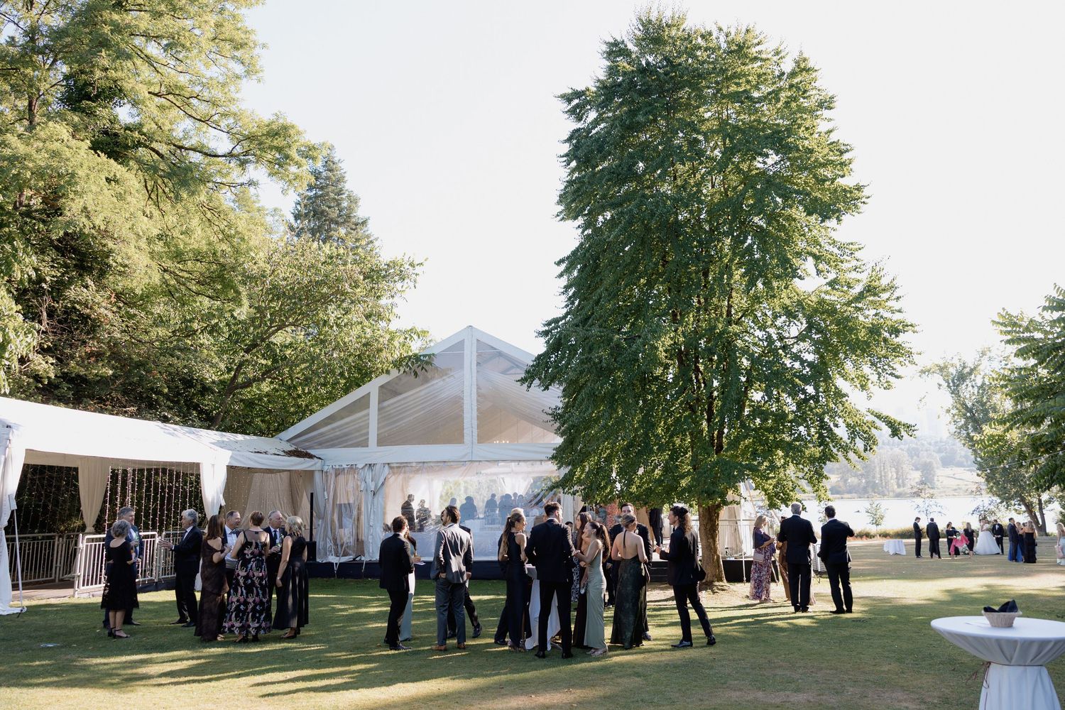 Wide shot of wedding guests gathering on a lawn with trees and a white tent in the distance Hurt House in Burnaby