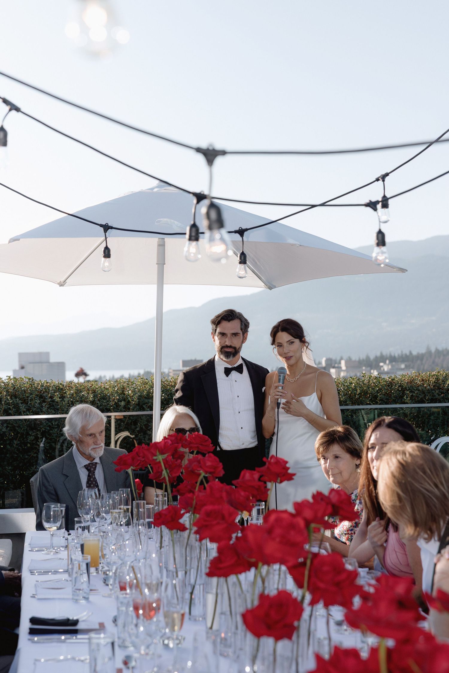 Elegant rooftop wedding reception with red floral centerpieces under string lights.