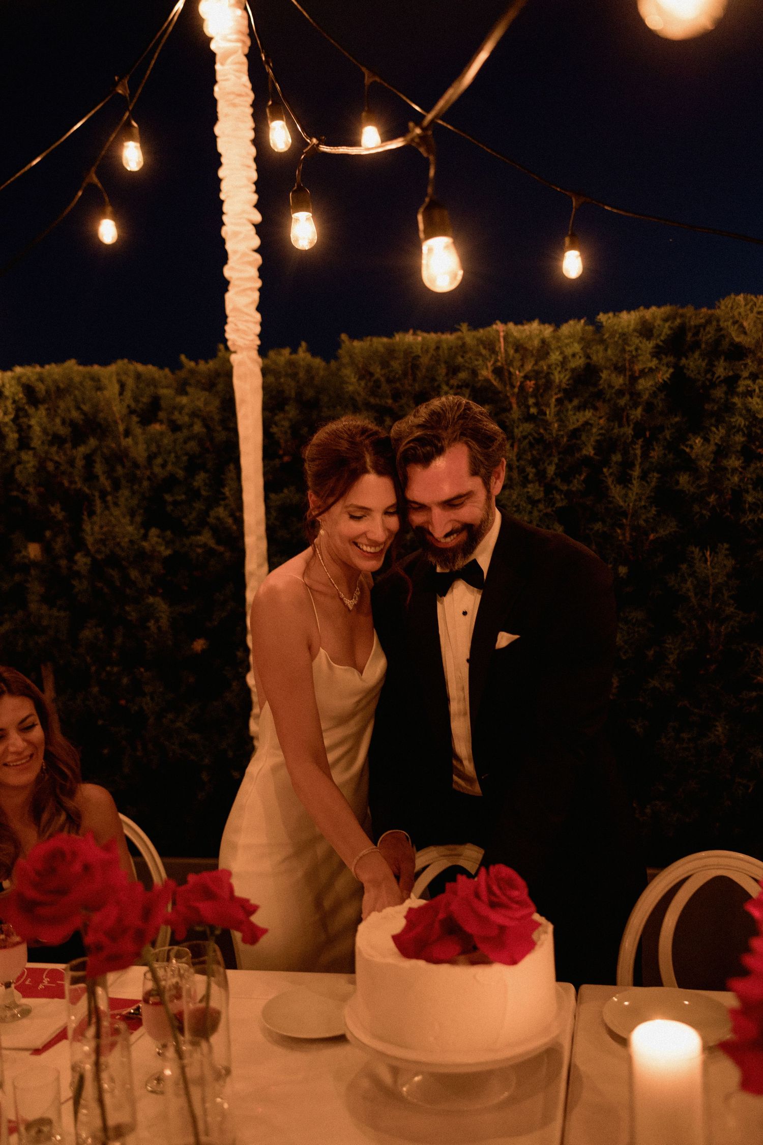 Romantic outdoor wedding reception with string lights and floral centerpieces and cutting cake
