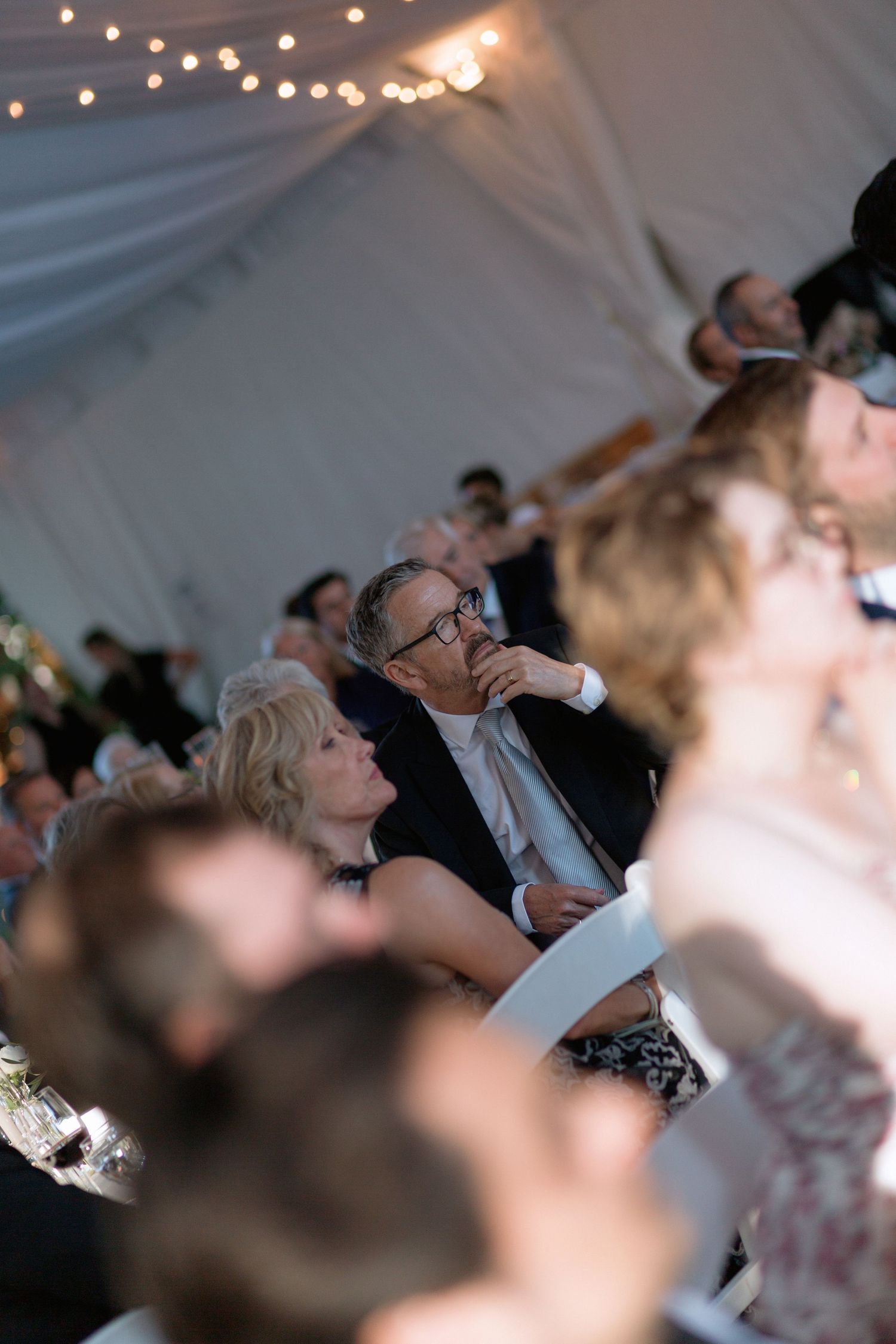 Wedding guests watching speeches during an evening reception under string lights.