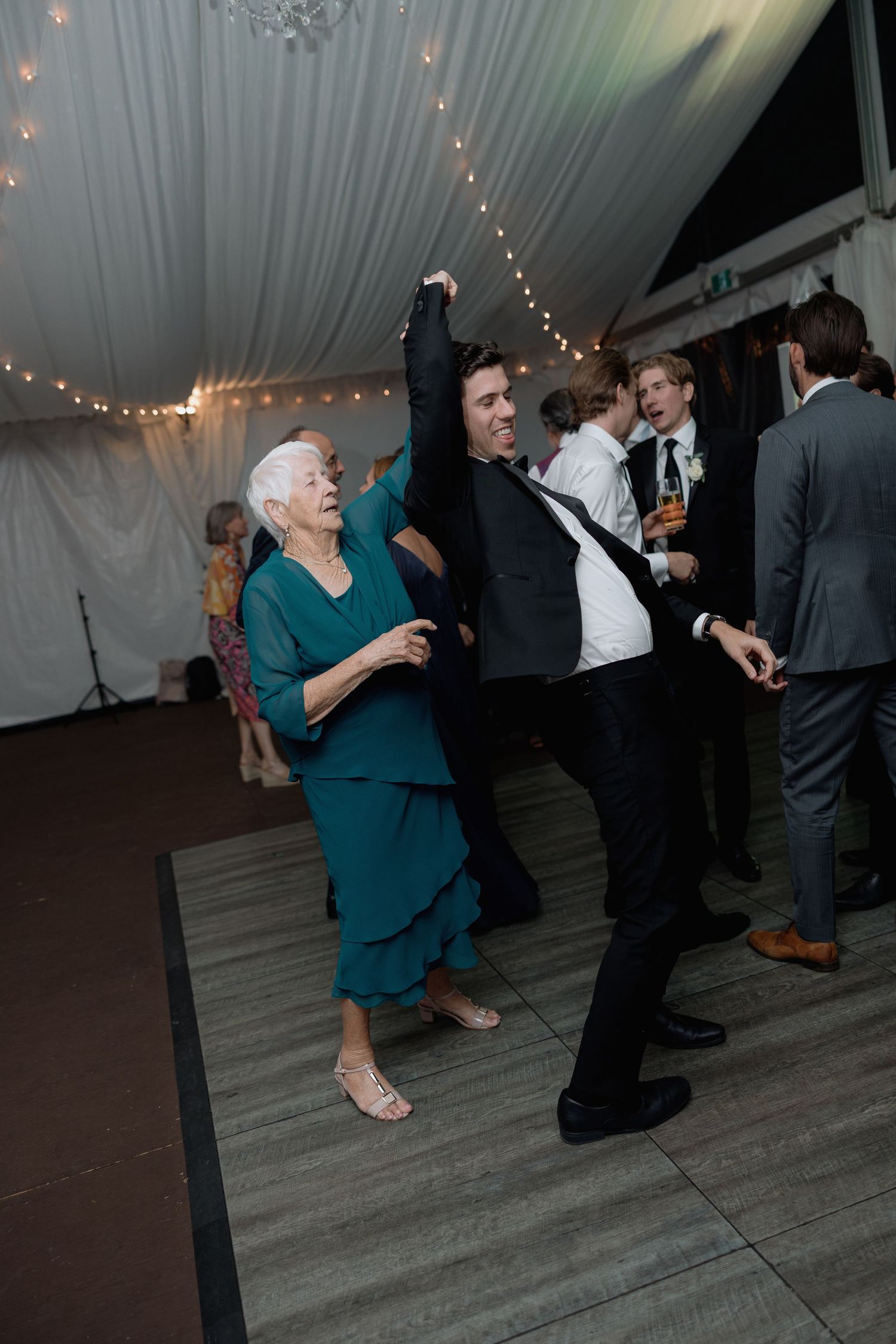 Joyful dancing at a wedding reception under string lights and white tent draping.
