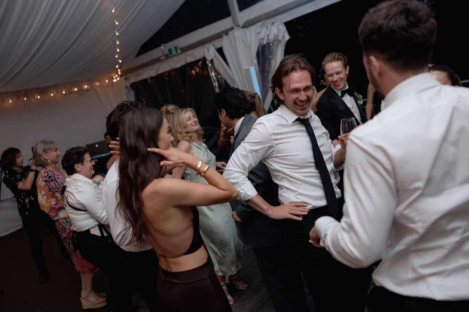 Energetic wedding guests dancing and celebrating at reception under white tent with string lights.