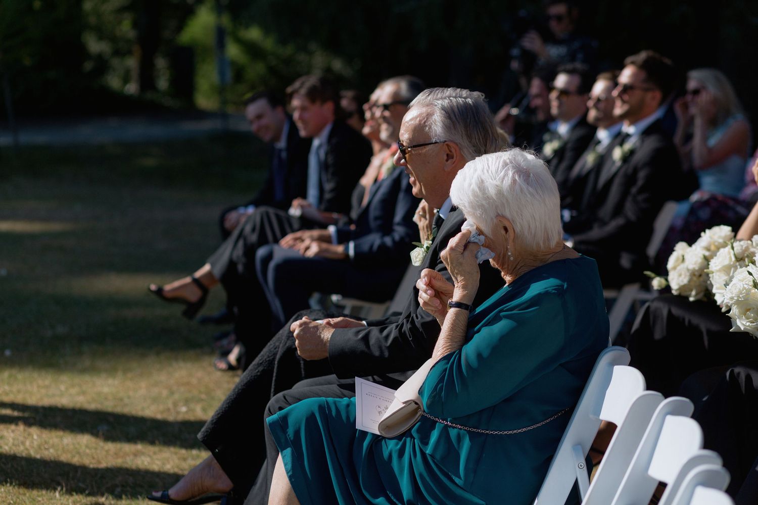 Wedding guests seated in white chairs watch the ceremony on a sunny outdoor day.