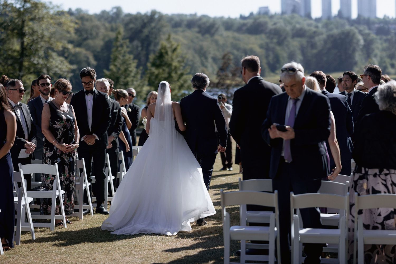 Wedding guests seated in white chairs at an outdoor ceremony with urban skyline in the distance.