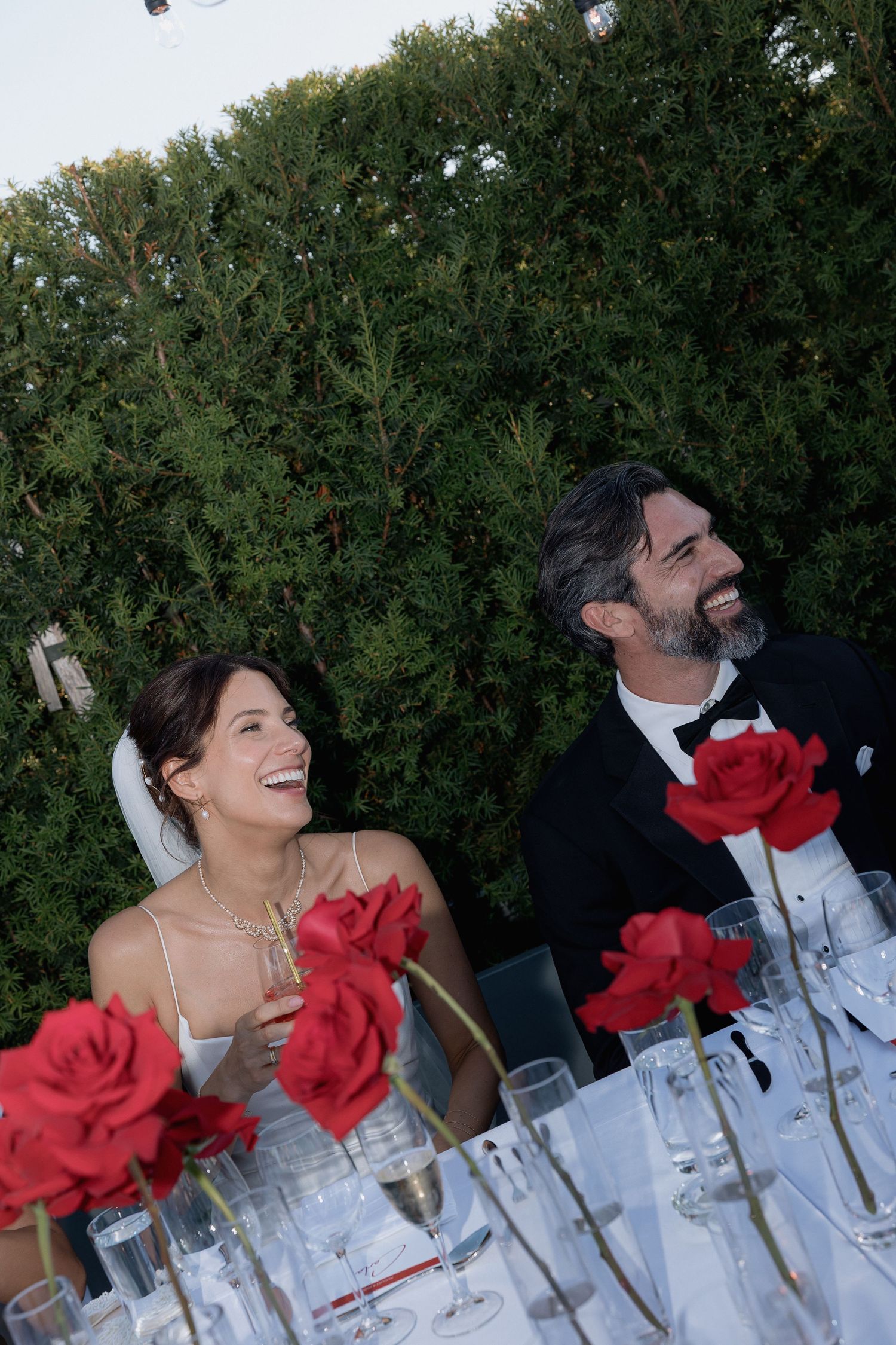 Wedding reception table decorated with red roses and wine glasses against green foliage.