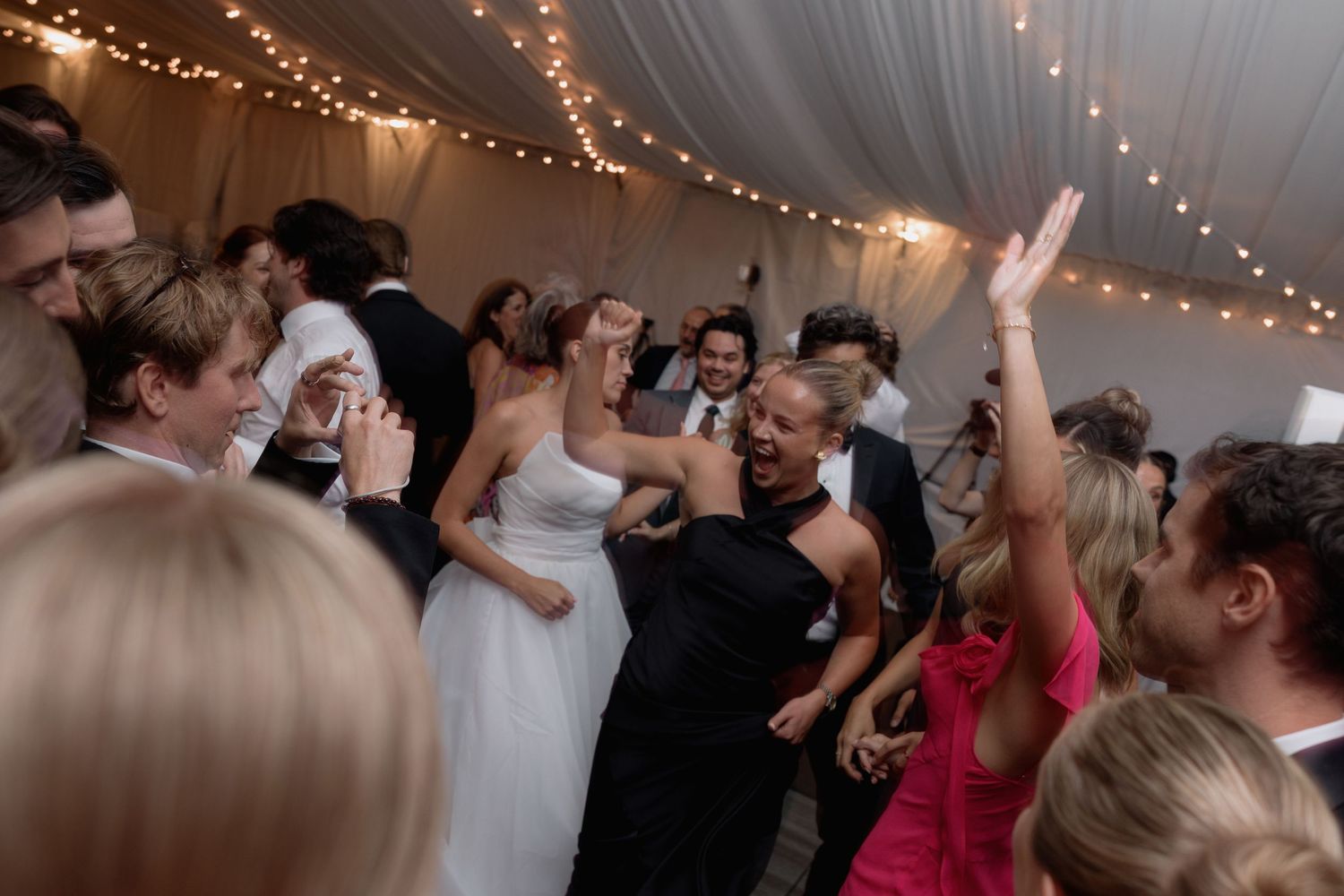 Lively dance floor scene with bride and wedding guests celebrating under string lights.