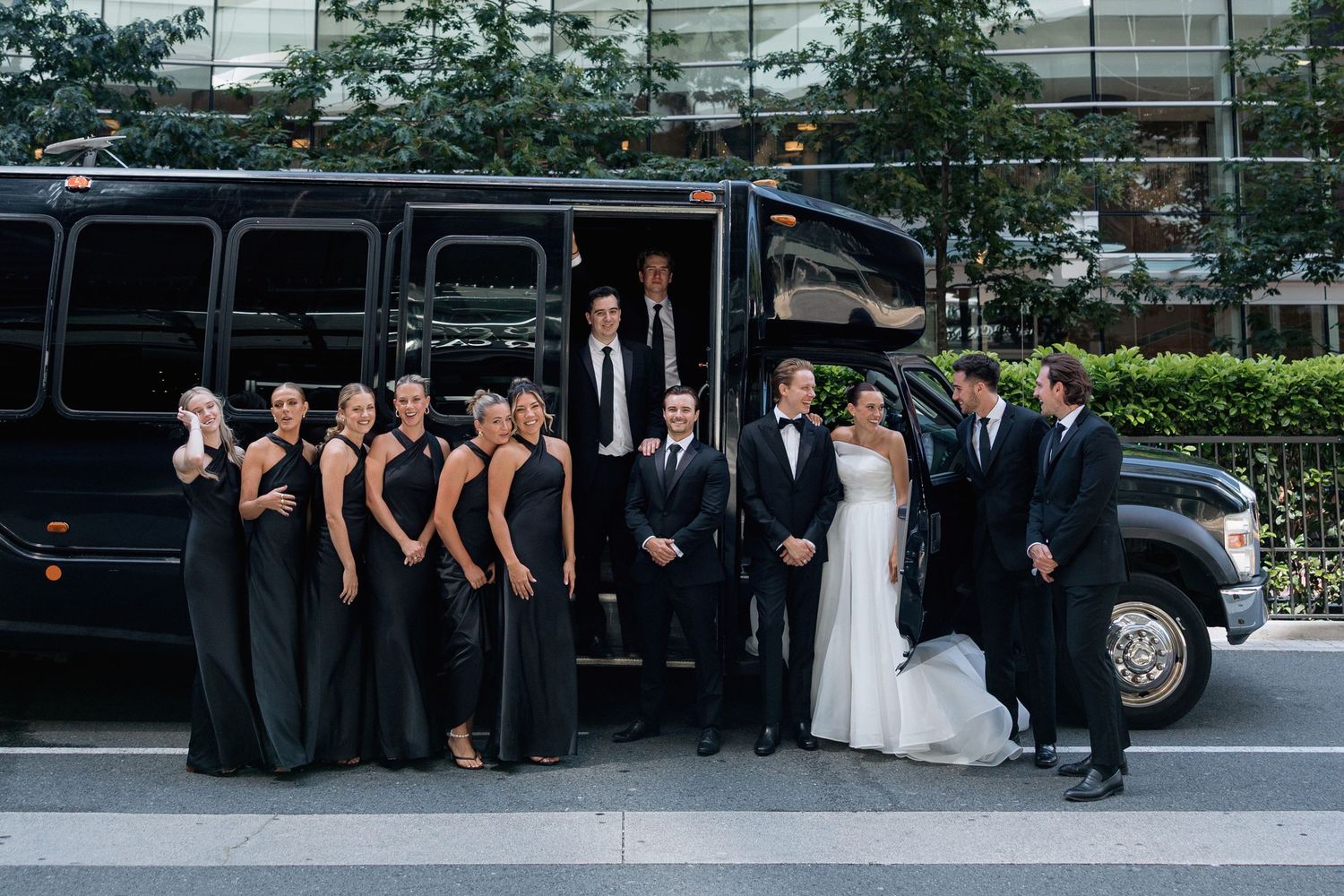 Wedding party members arranged near black limousine with urban architecture backdrop.