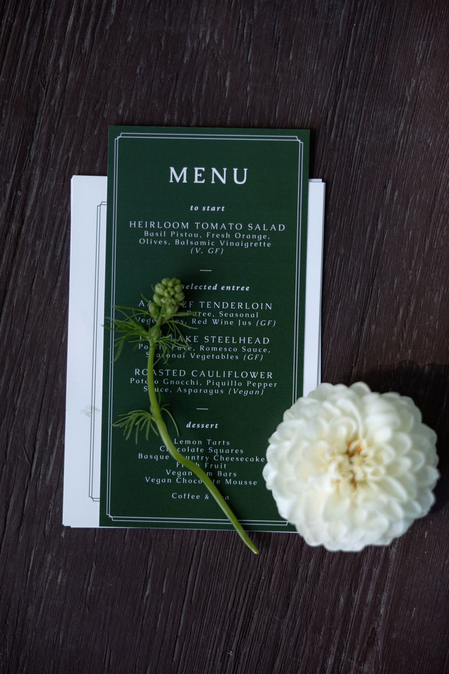 Elegant green wedding menu card with white flower and greenery sprig on dark wooden surface.