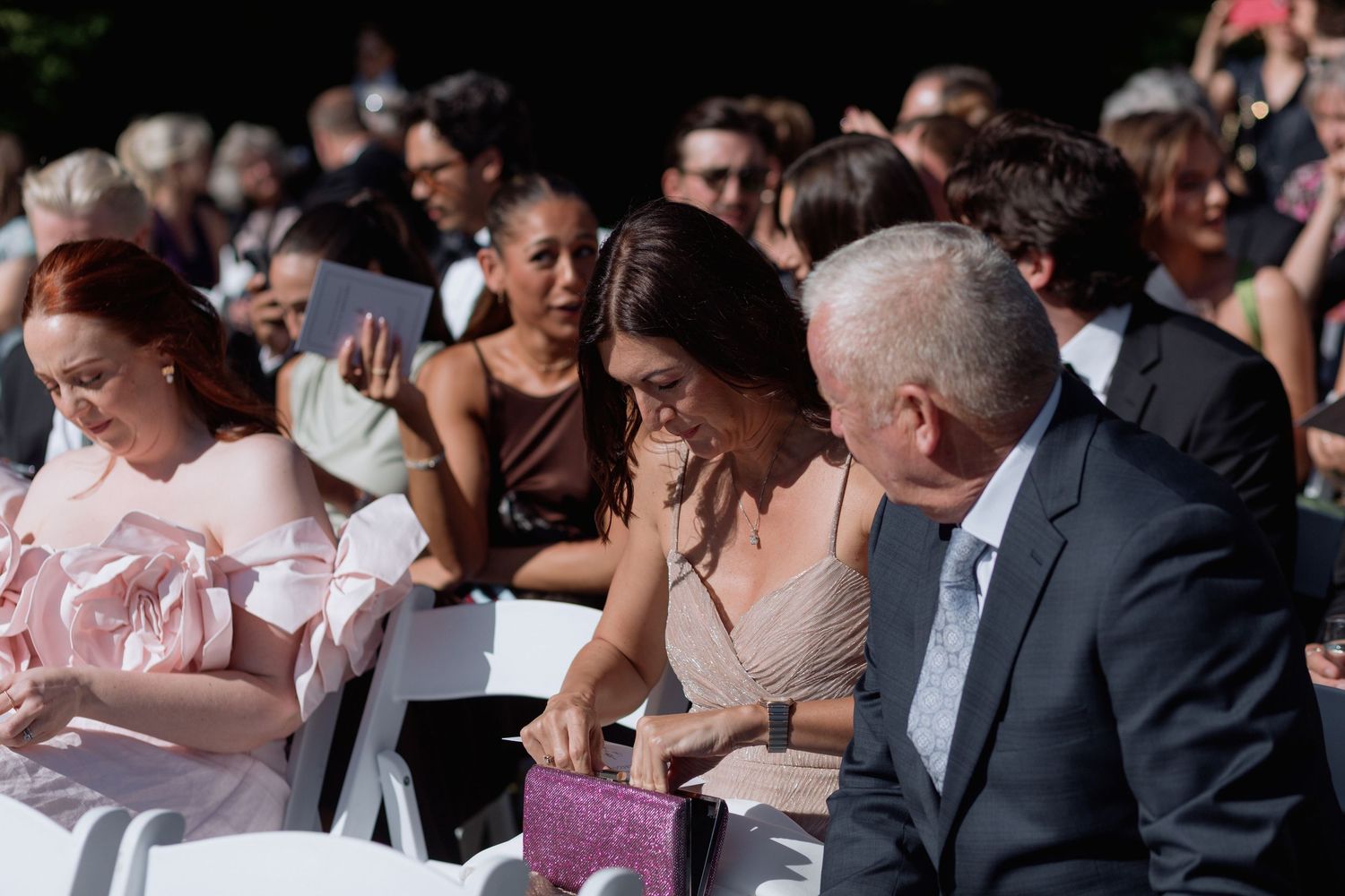Wedding attendees in formal wear watching ceremony from white chairs.