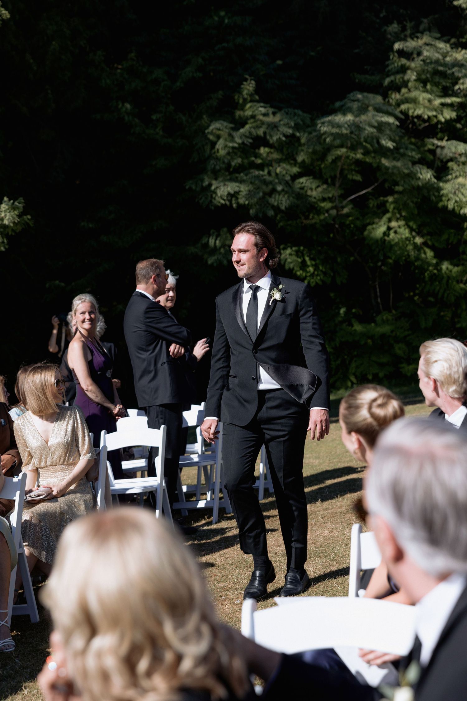 Wedding guests seated in white chairs at an outdoor ceremony surrounded by trees.