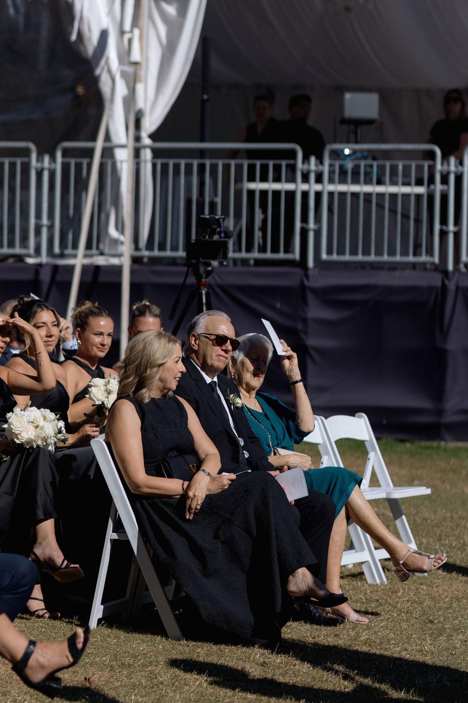 Wedding guests seated in formal attire on white chairs during an outdoor ceremony.