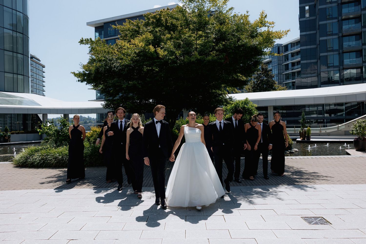 Panoramic shot of wedding party gathered on urban plaza with modern buildings and trees at Marriott Hotel in Vancouver