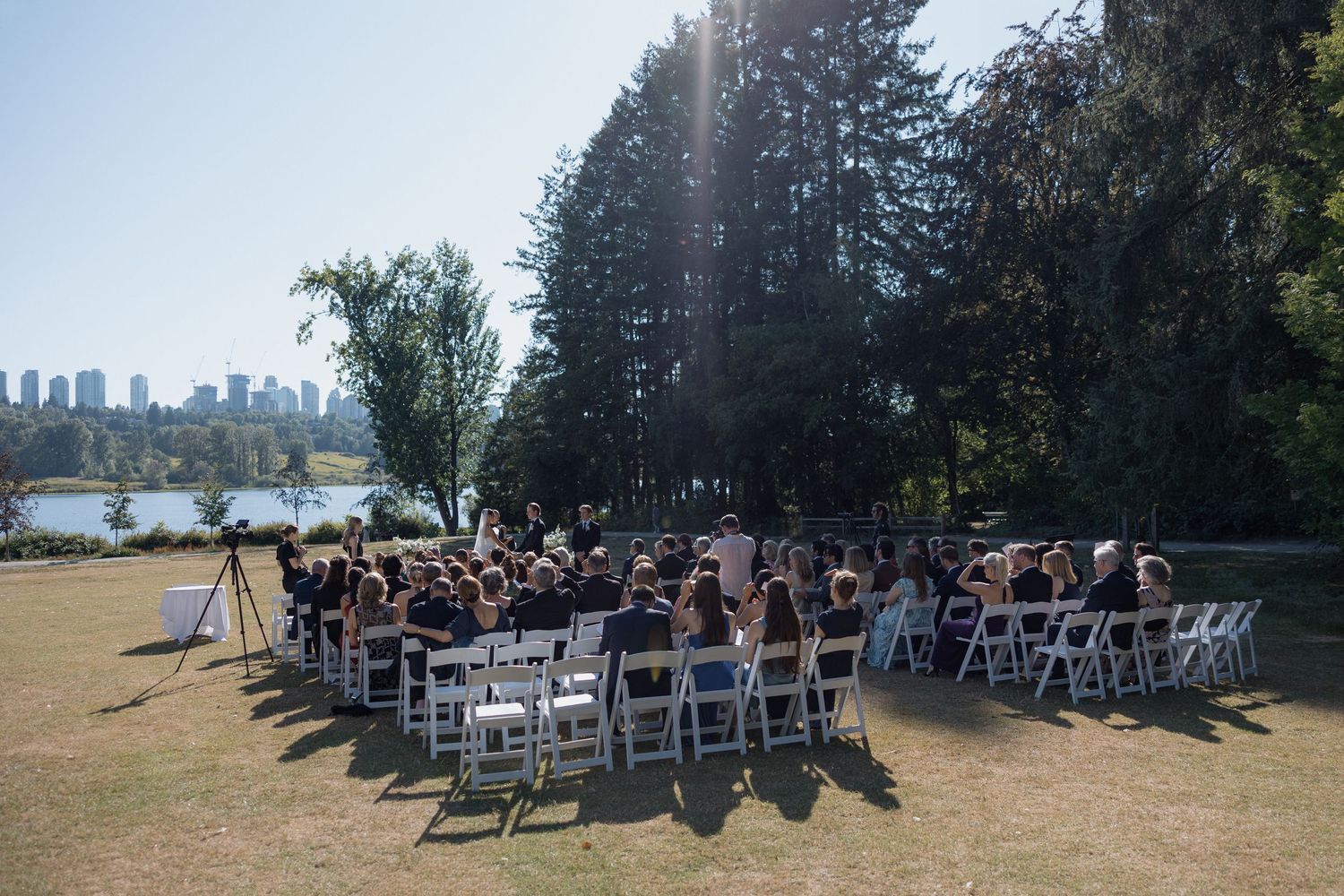 Wide angle shot of wedding venue with white chairs arranged on grass near trees and with a view on Deer Lake at Hurt House in Burnaby