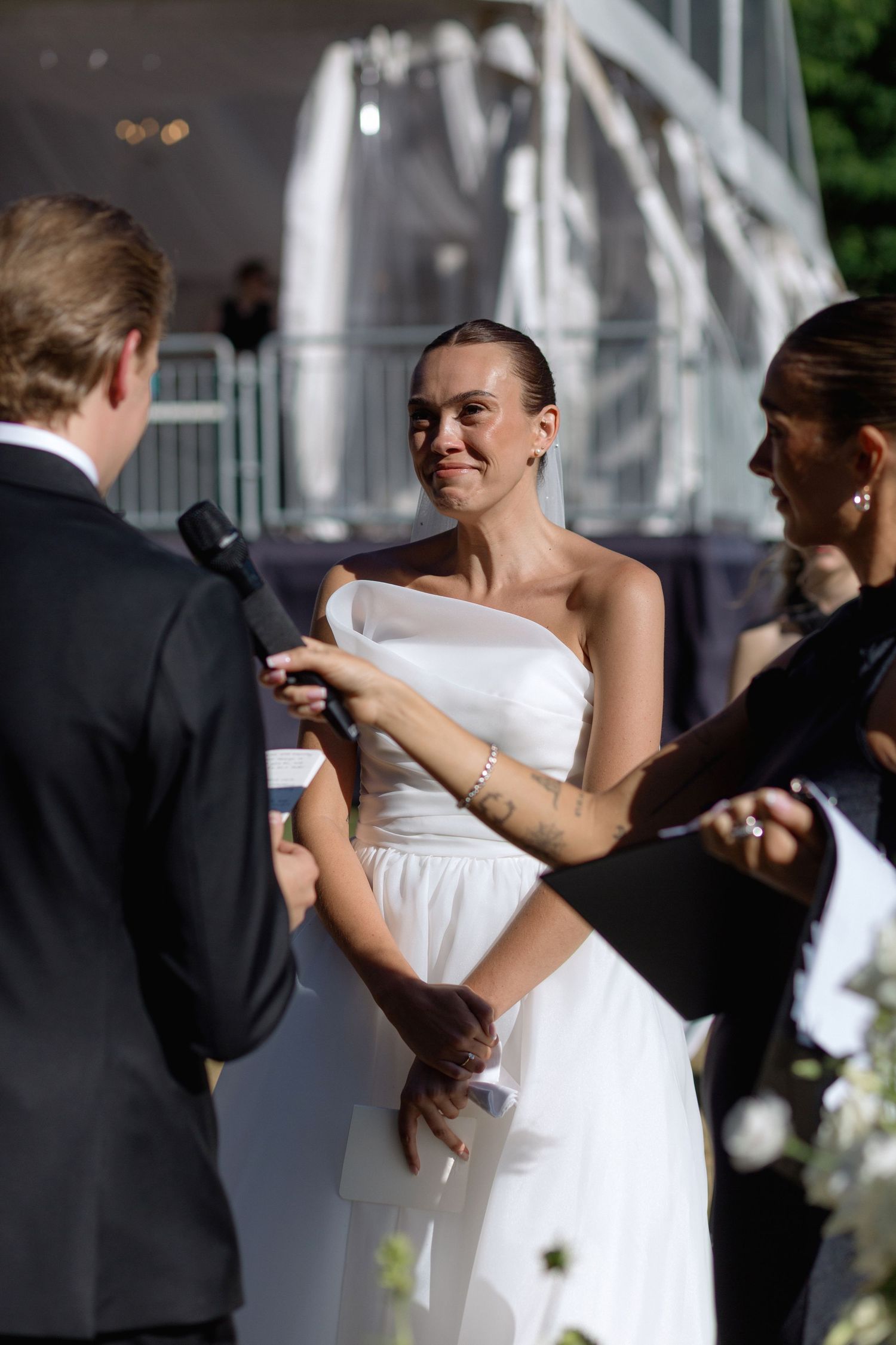 A bride in a white gown exchanges vows during an outdoor ceremony beneath a white gazebo.