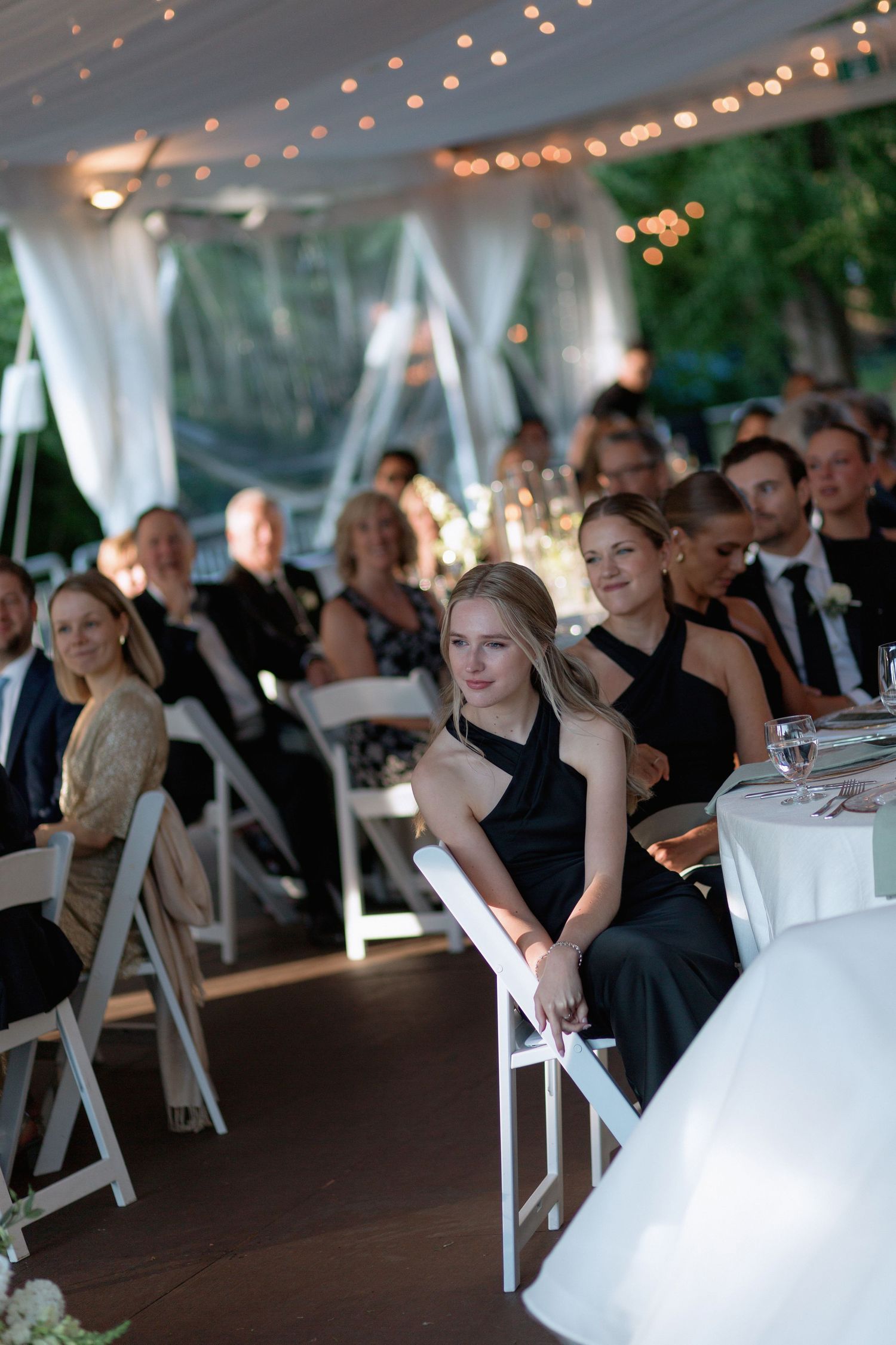 Formal wedding reception with guests seated at white-clothed tables under string lights in an outdoor tent.