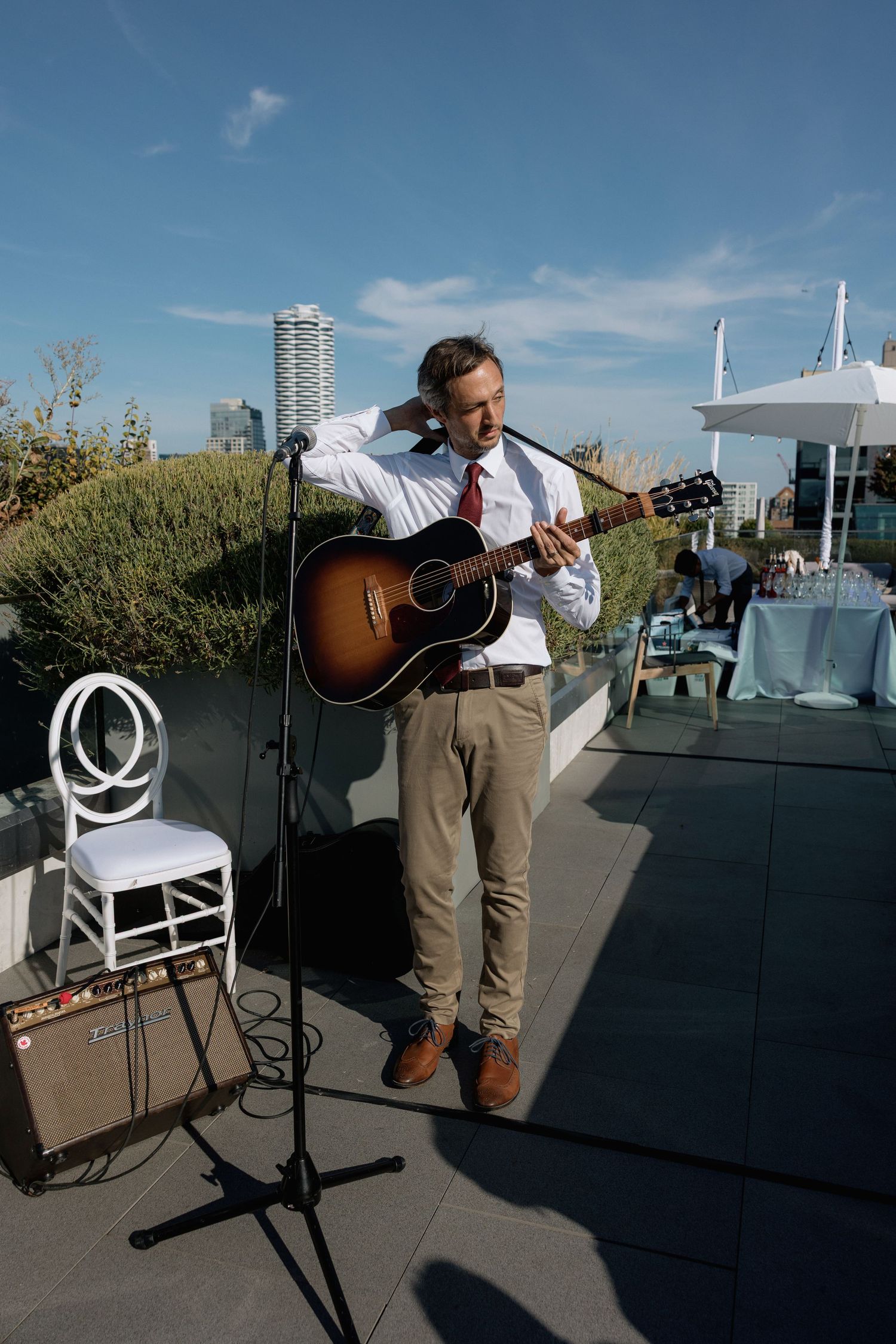 Musician playing acoustic guitar on a rooftop terrace against blue sky and city backdrop.