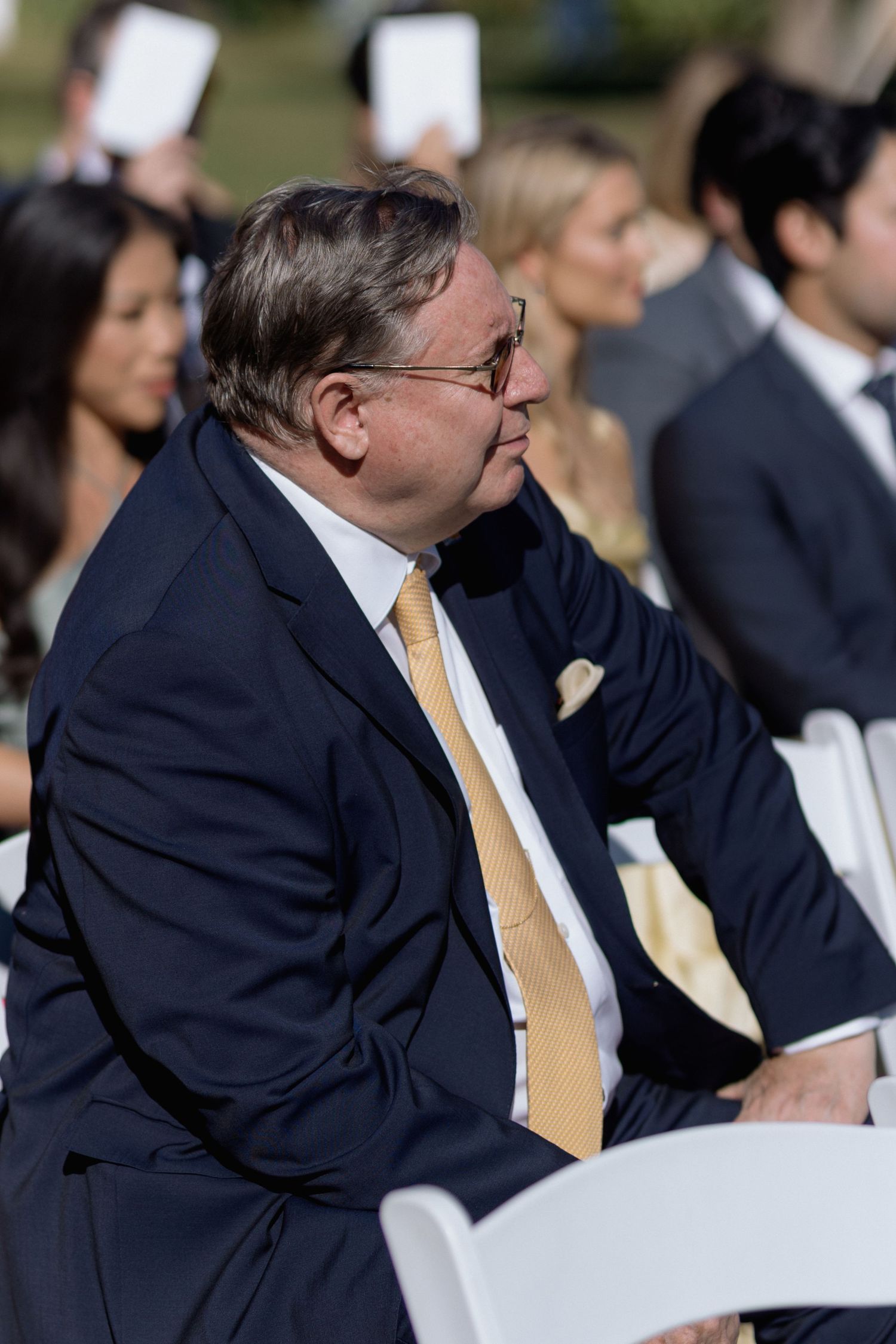 Wedding guest in navy suit and yellow tie seated outdoors during ceremony.
