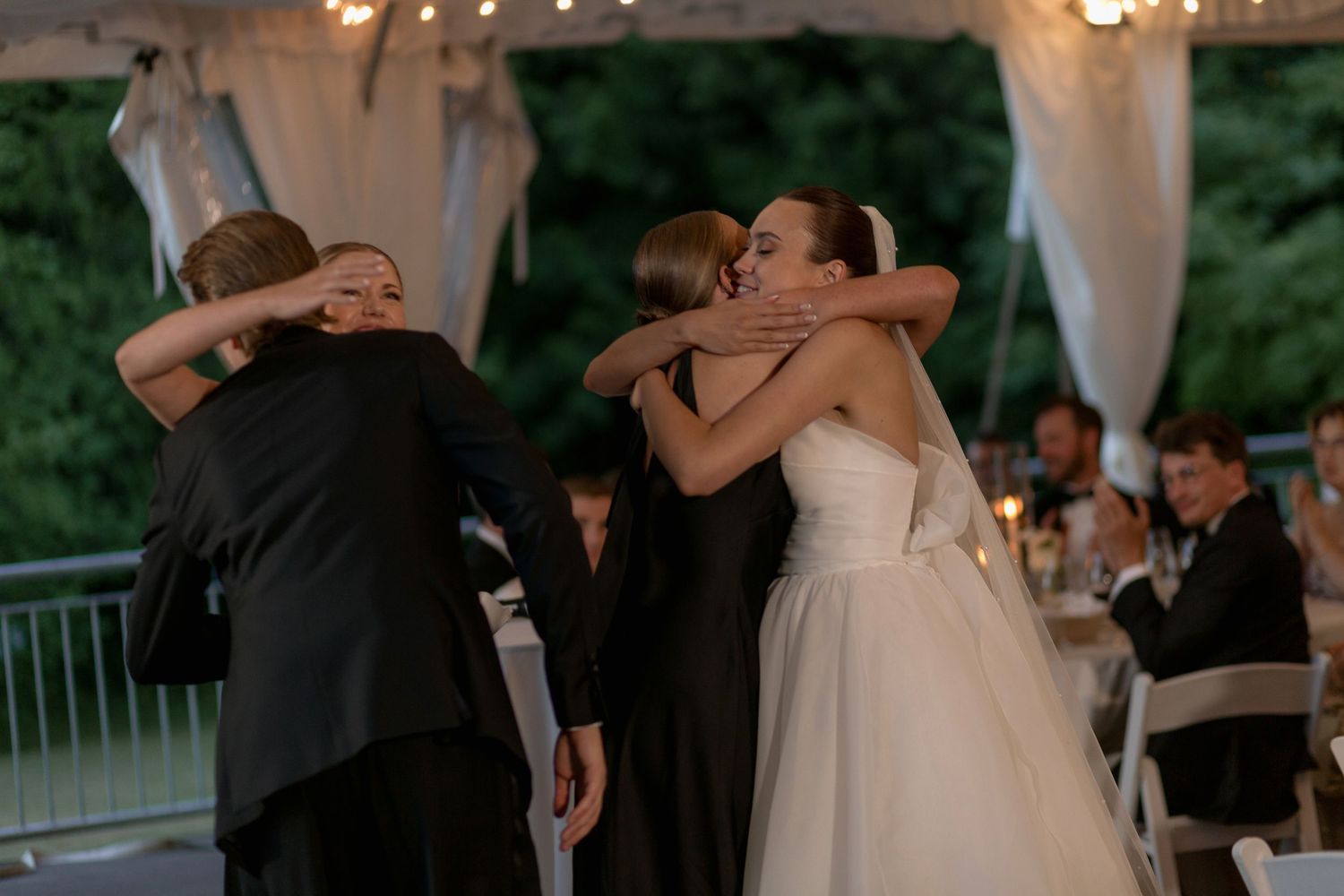Emotional group embrace during wedding reception under white tent and string lights.