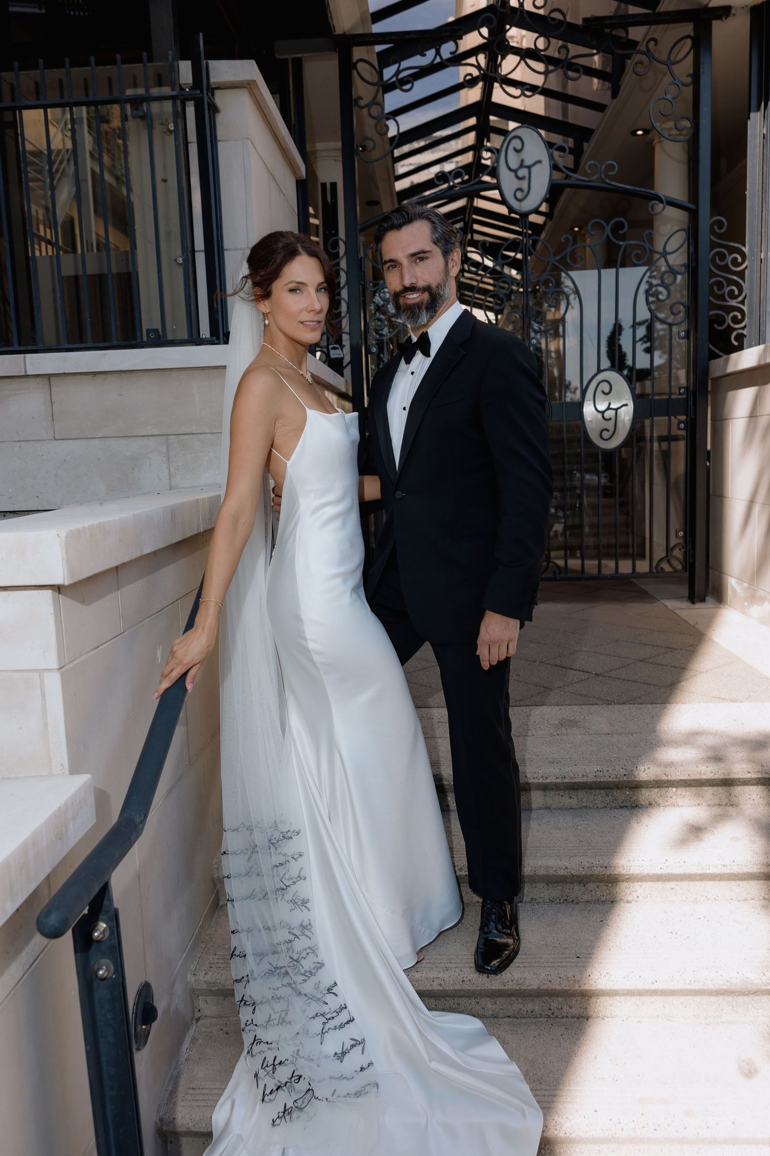 A bride in a flowing white gown and groom in black suit walk together under a covered walkway with ornate railings.