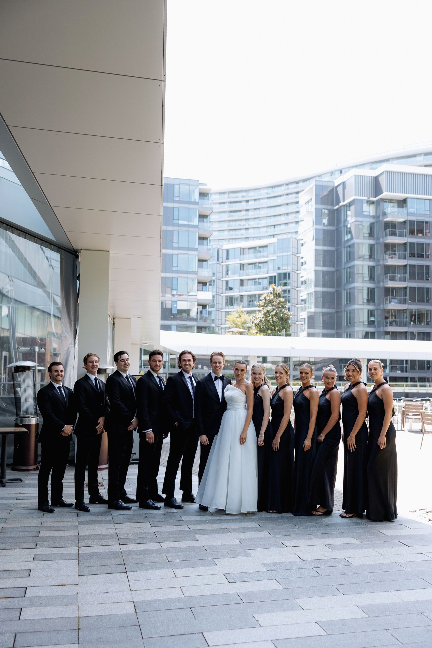 Wedding party in formal attire poses for group photos on a modern terrace with city buildings in background at Marriott Hotel in Vancouver