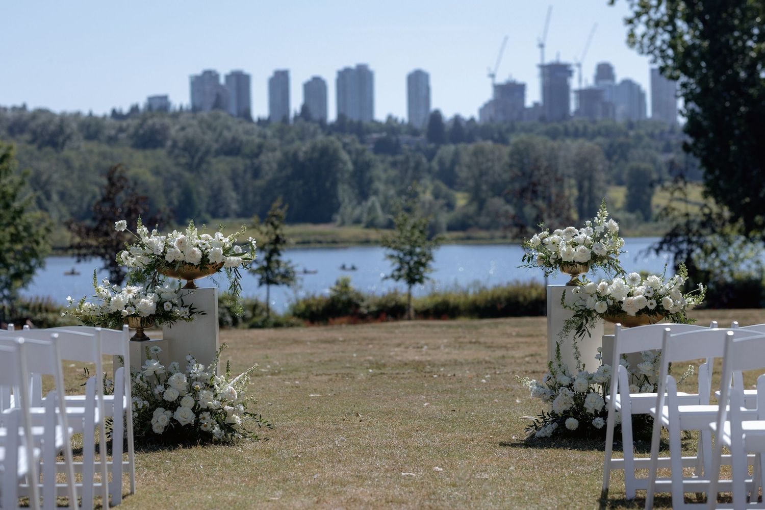 White ceremony chairs and floral arrangements line an outdoor aisle with a cityscape backdrop and Deer Lake at Hurt House in Burnaby