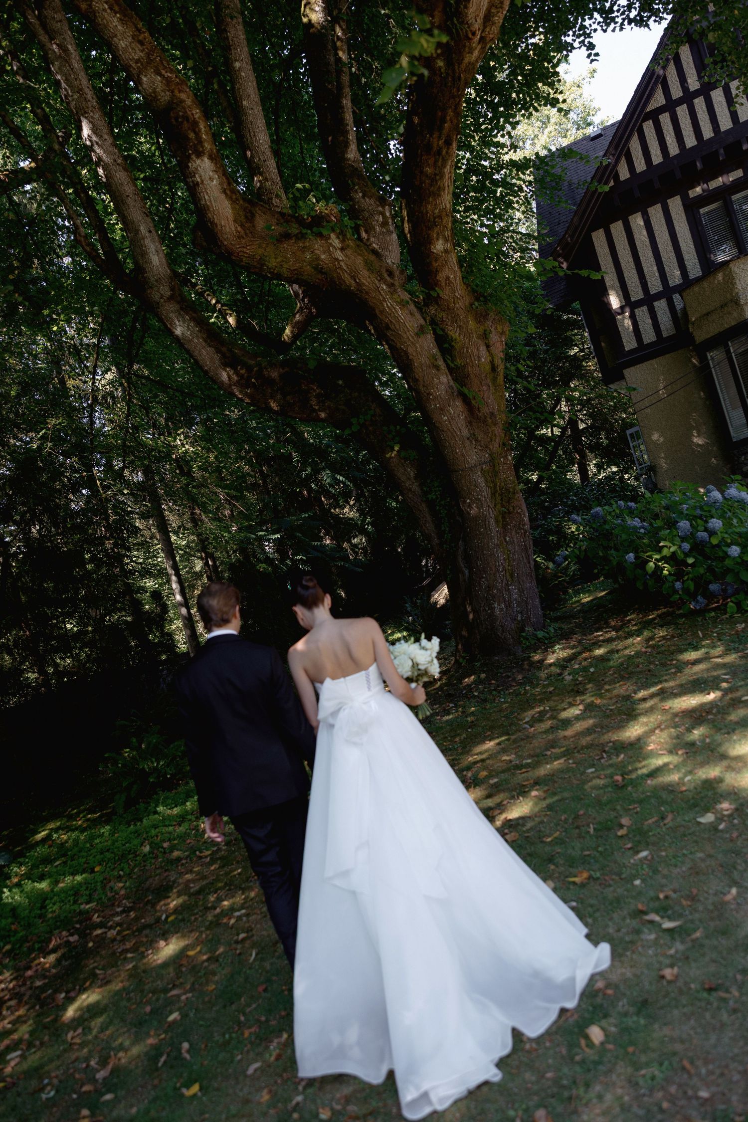 Couple in wedding attire walking together through a lush garden path with trees overhead.