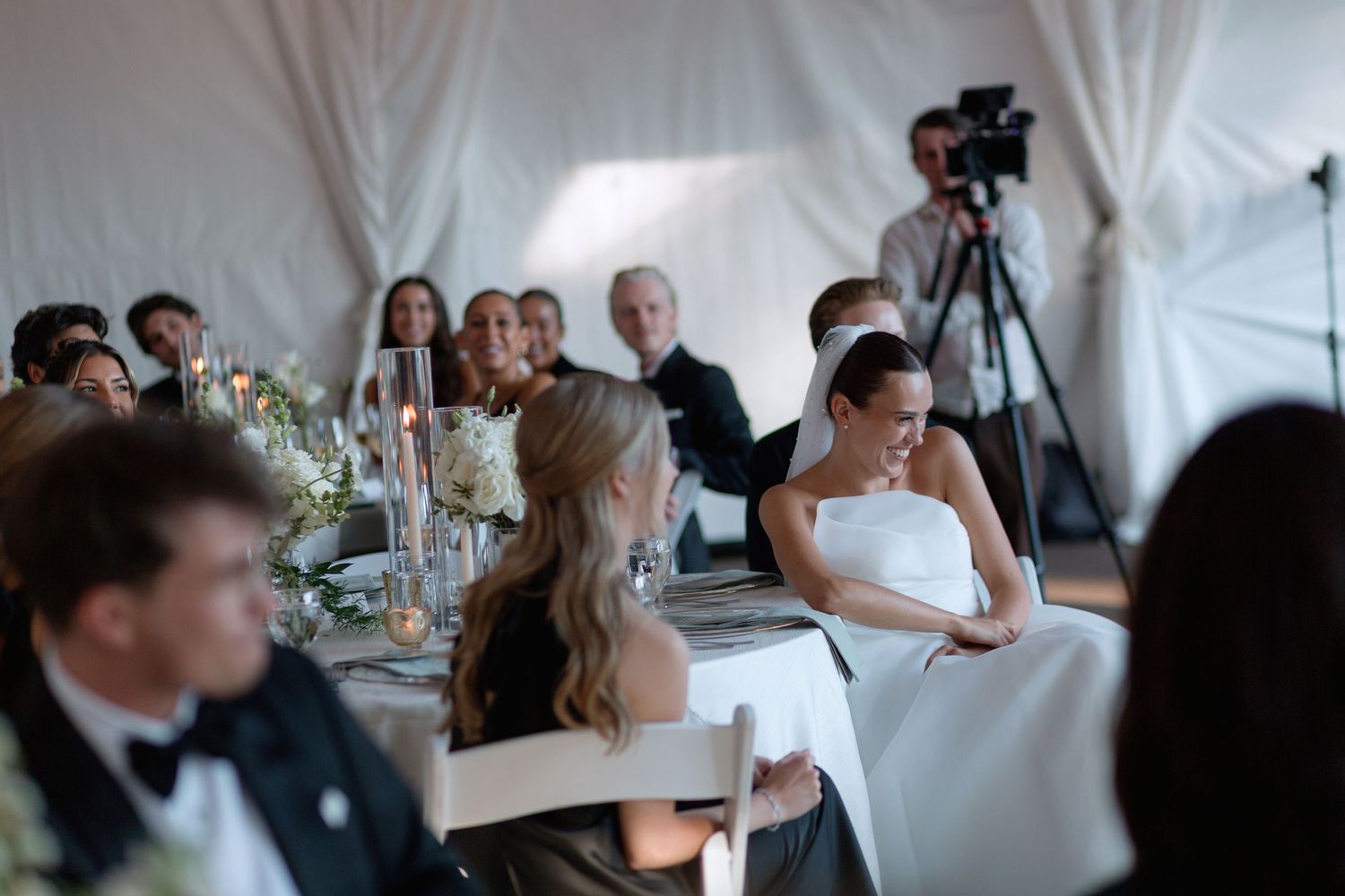 Wedding guests seated at round tables during reception dinner under white tent.