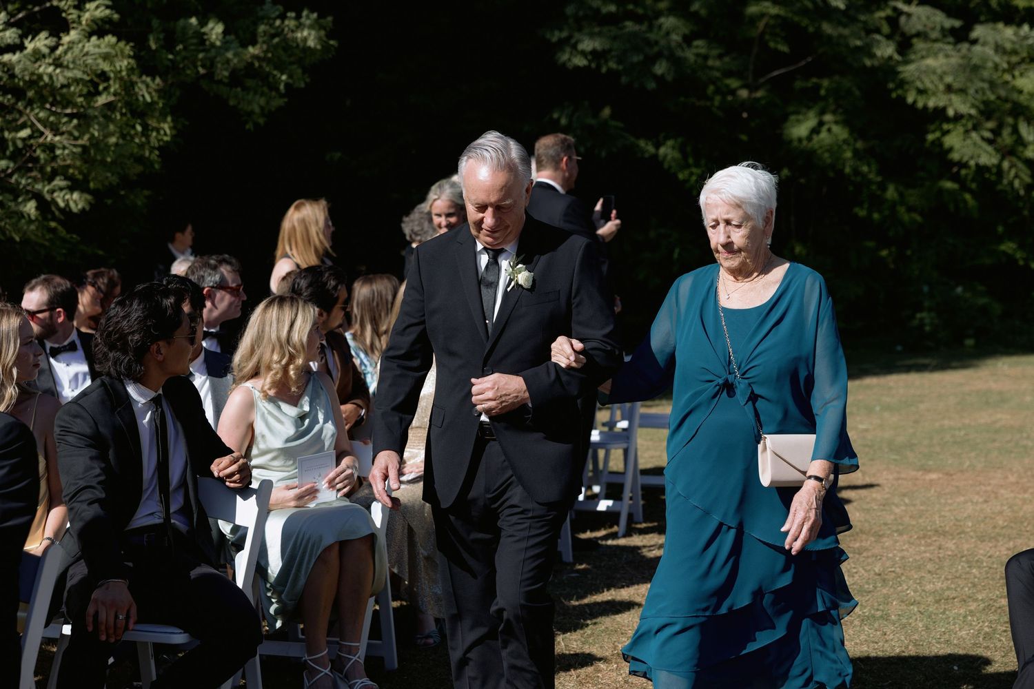 Attendees in formal wear and teal dresses gather at an outdoor wedding venue.