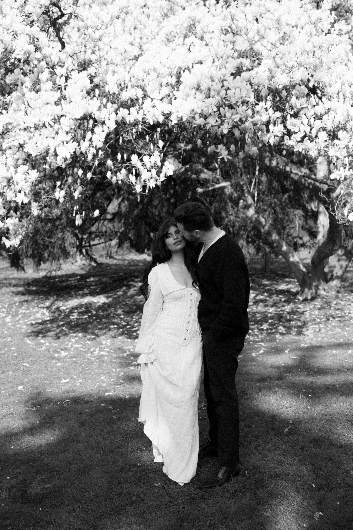 A romantic black and white wedding photo showing a couple embracing under a blossoming tree in a garden setting.