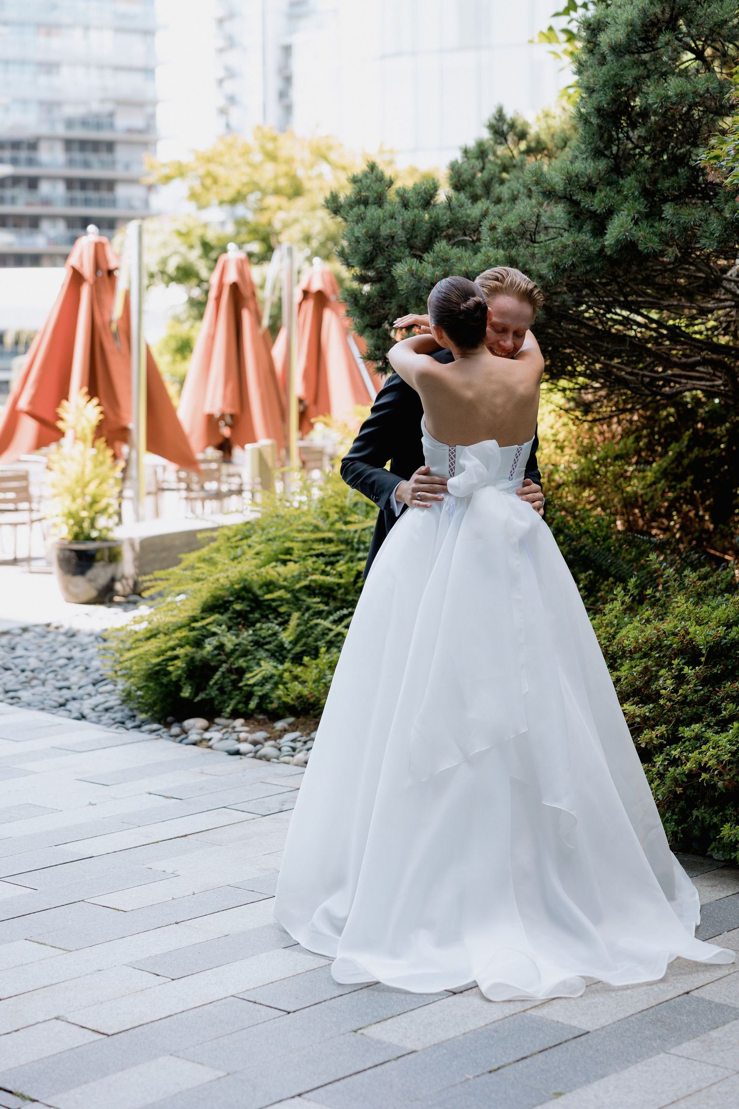 A tender embrace between newlyweds with orange patio umbrellas and modern architecture in the background at Marriott Hotel in Vancouver
