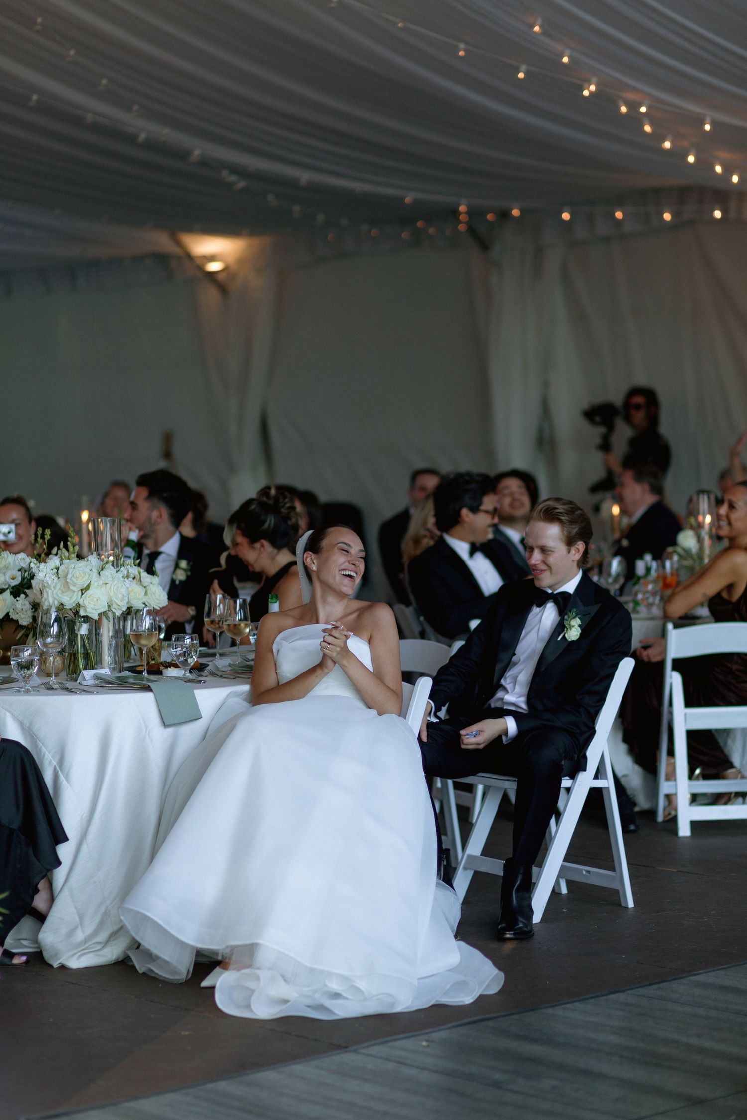 Wedding reception guests laughing and celebrating at tables under a draped white tent.