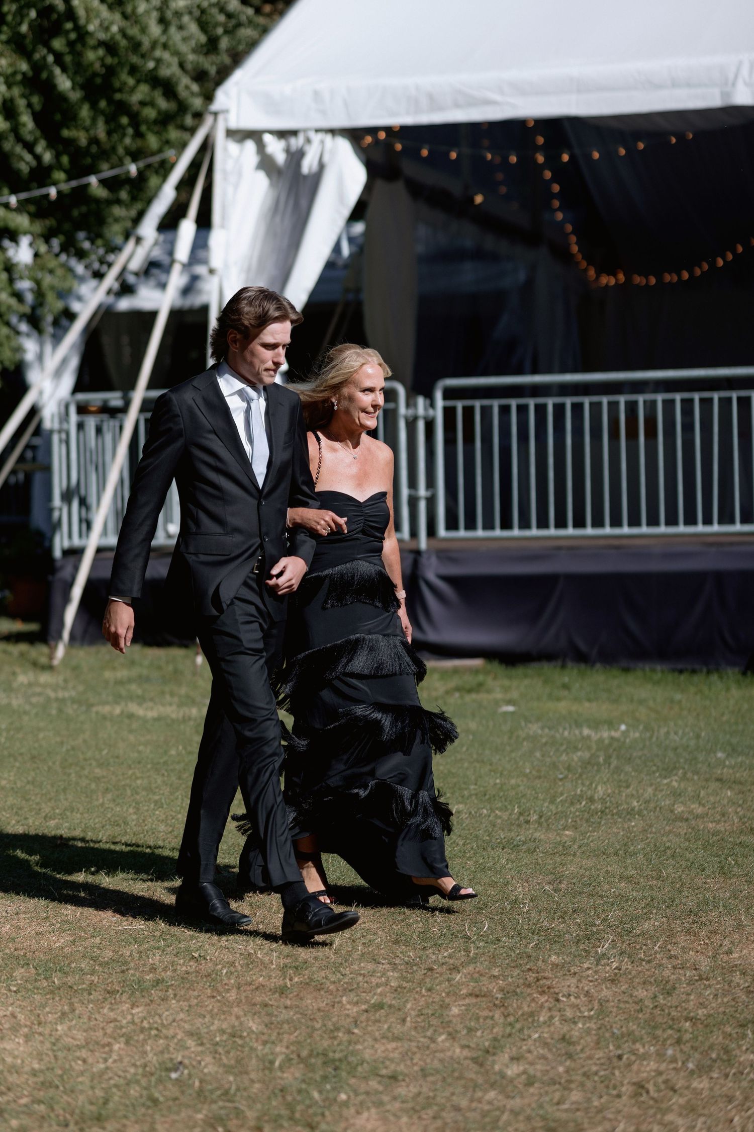 Formal couple walking together near white wedding tent with string lights.