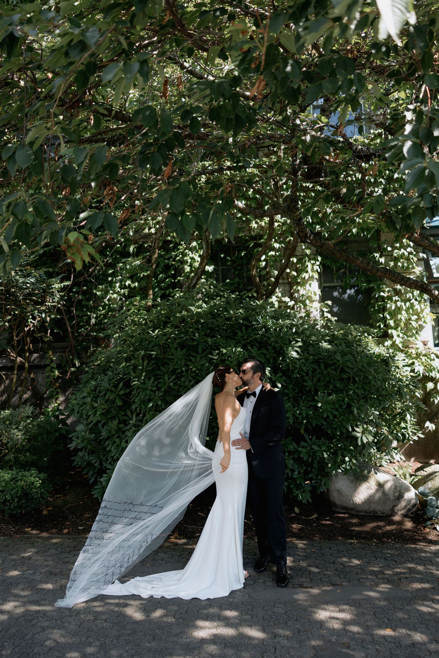 A romantic wedding moment captured outdoors with a flowing veil against a backdrop of green trees.