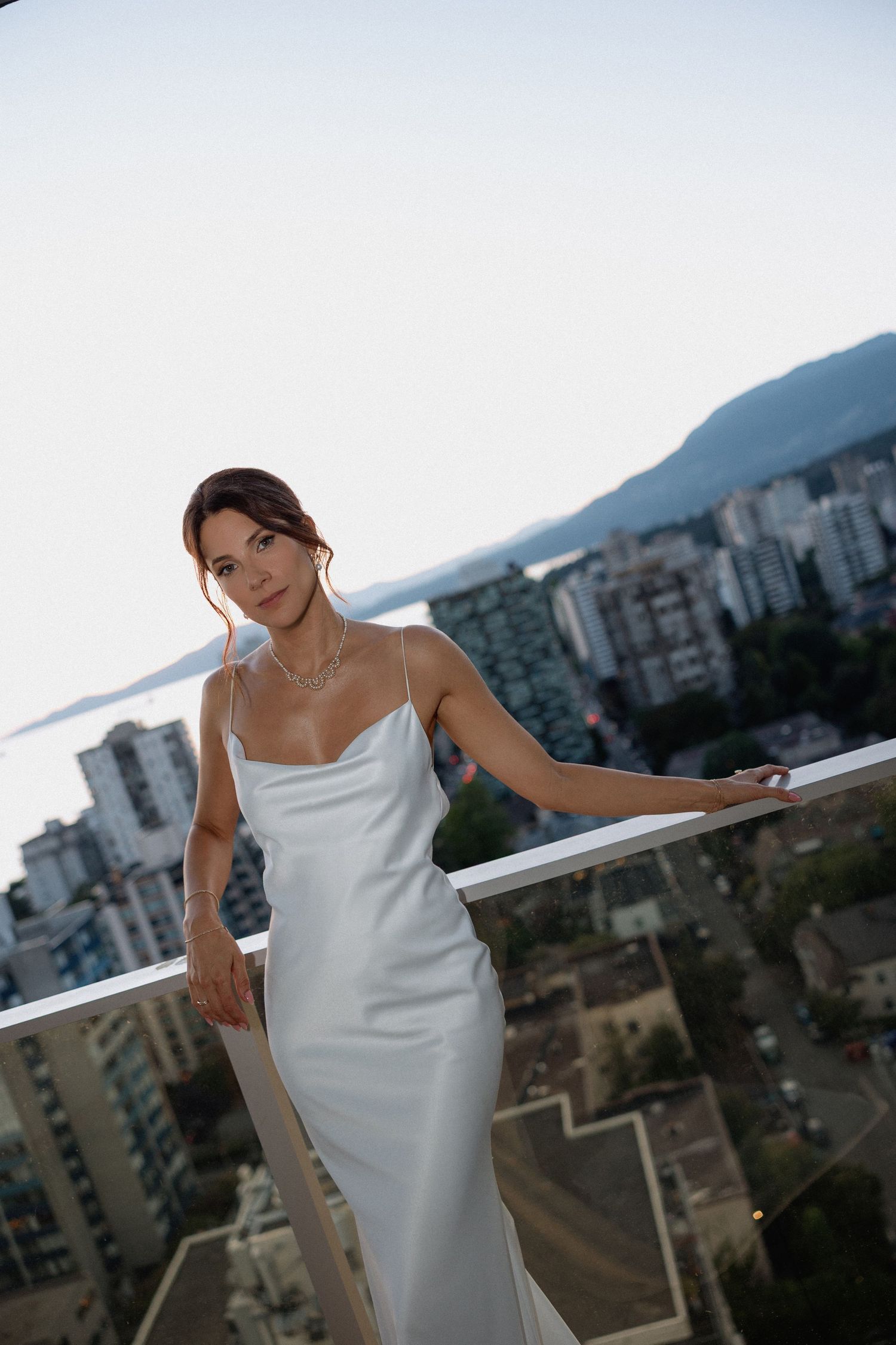 Person in white dress poses on balcony with cityscape and mountains in background.