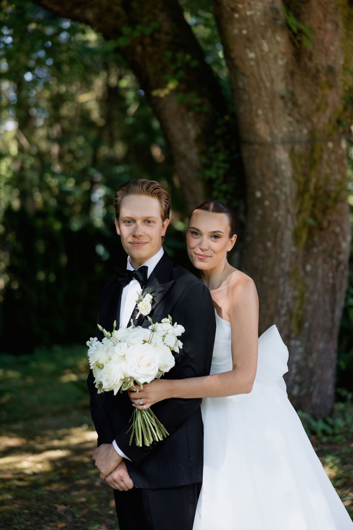 Newlyweds pose together in outdoor garden setting with white floral bouquet.