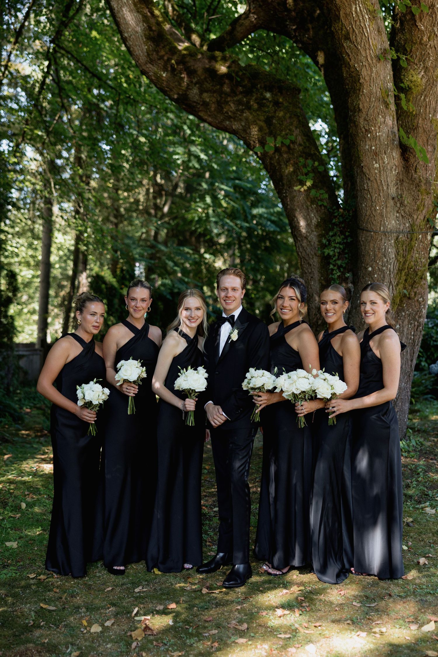 Wedding party in elegant black attire poses under a large tree with white bouquets.