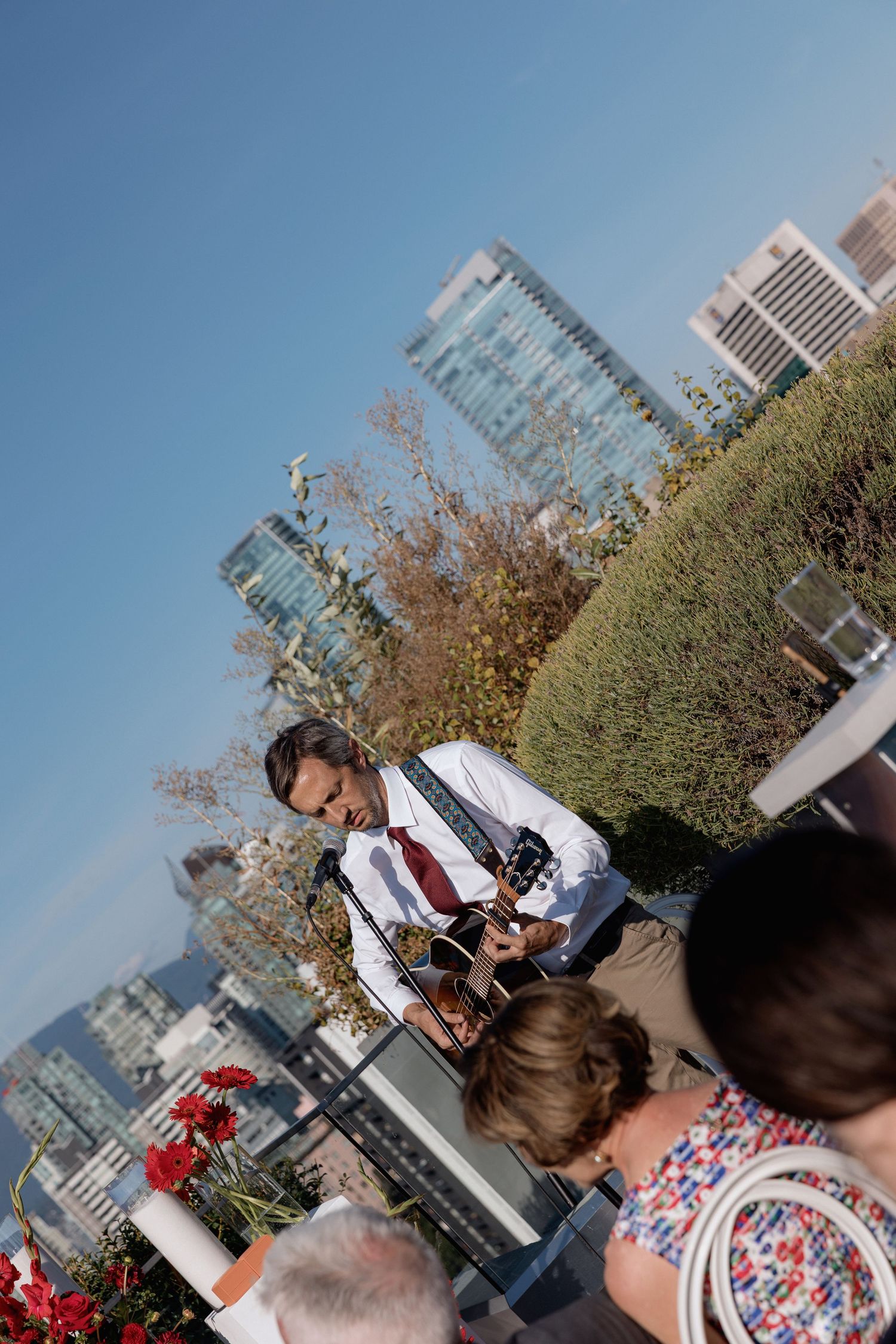 Outdoor rooftop gathering with string lights and red flowers on tables overlooking a city skyline.